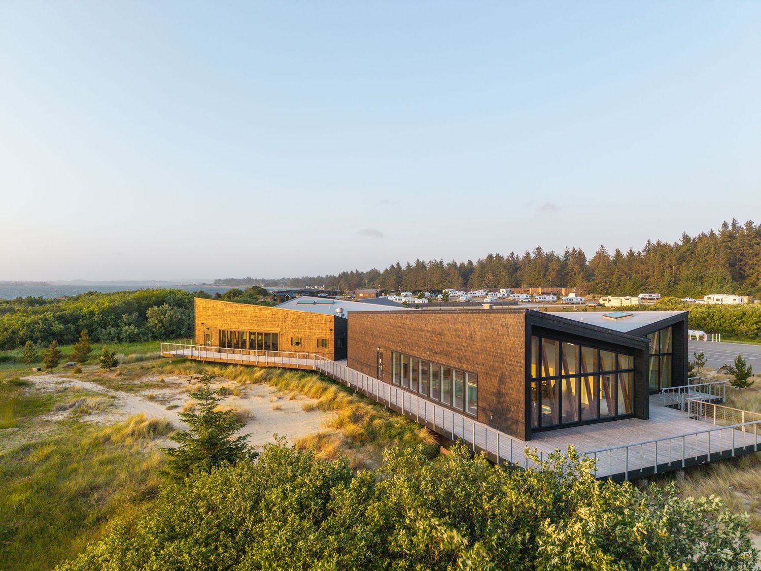 Aerial view of a modern wooden building surrounded by sand dunes and pine forest under a clear blue sky.