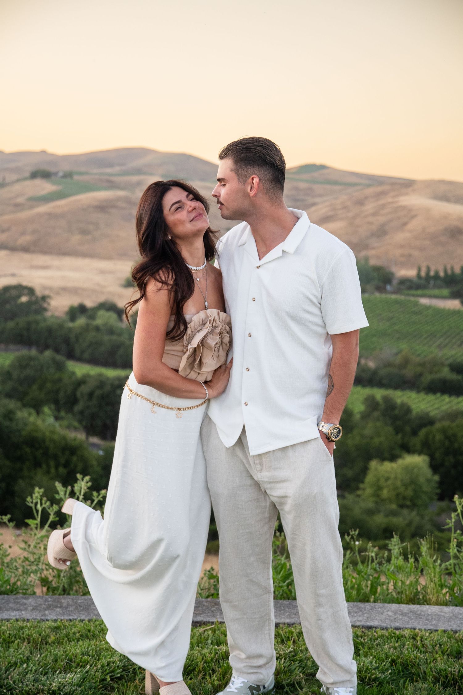 Romantic couple in white outfits pose against rolling hills at sunset.