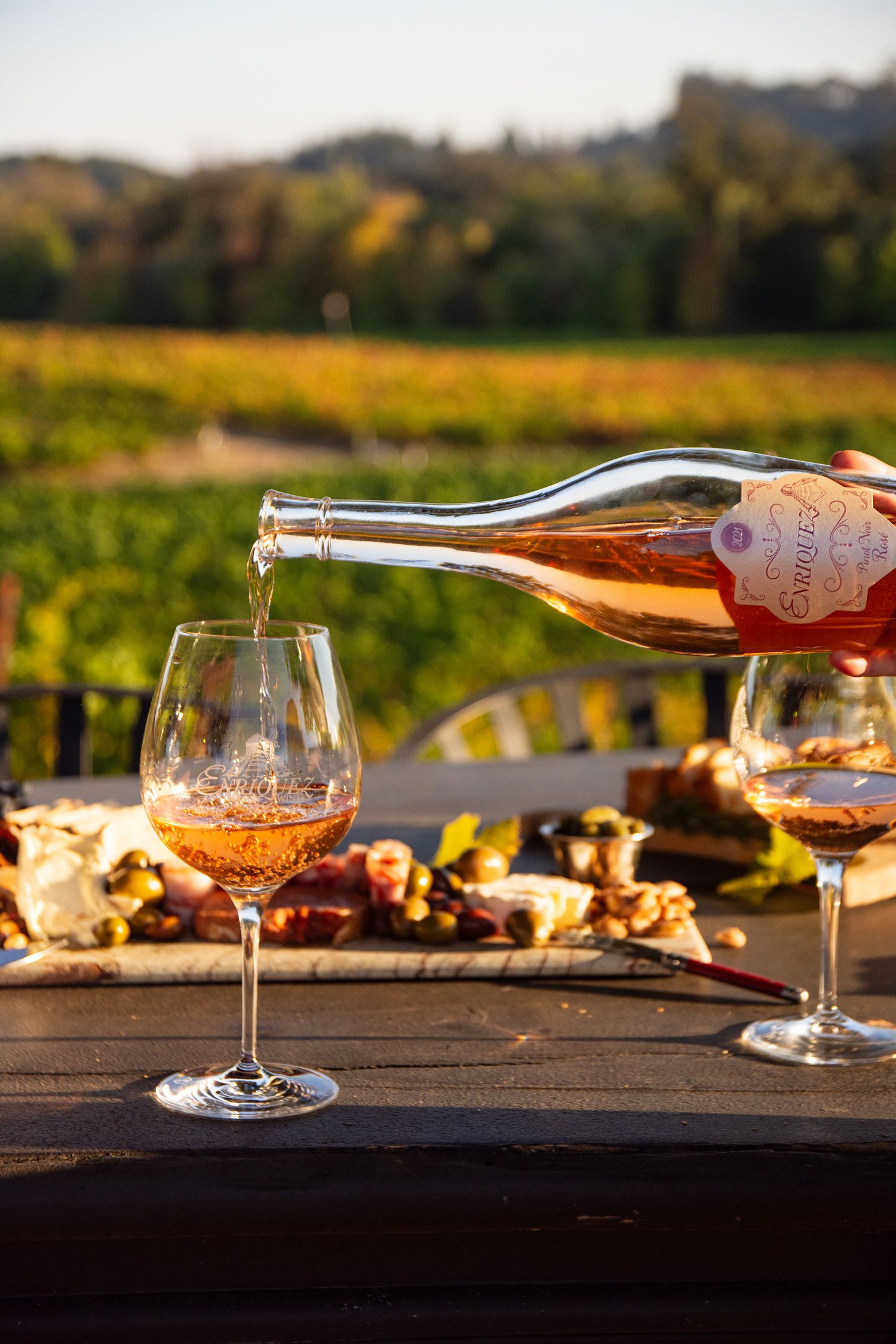 Rosé wine being poured into a glass on a wooden table with a charcuterie board and vineyard views.