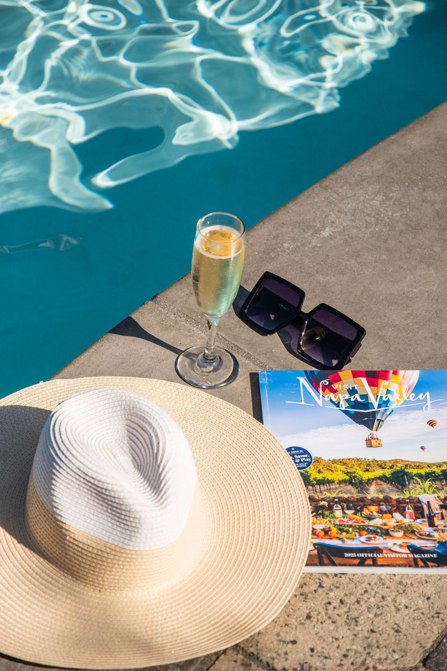 Poolside scene with sunhat, champagne glass, and Nevada travel brochure beside turquoise water.