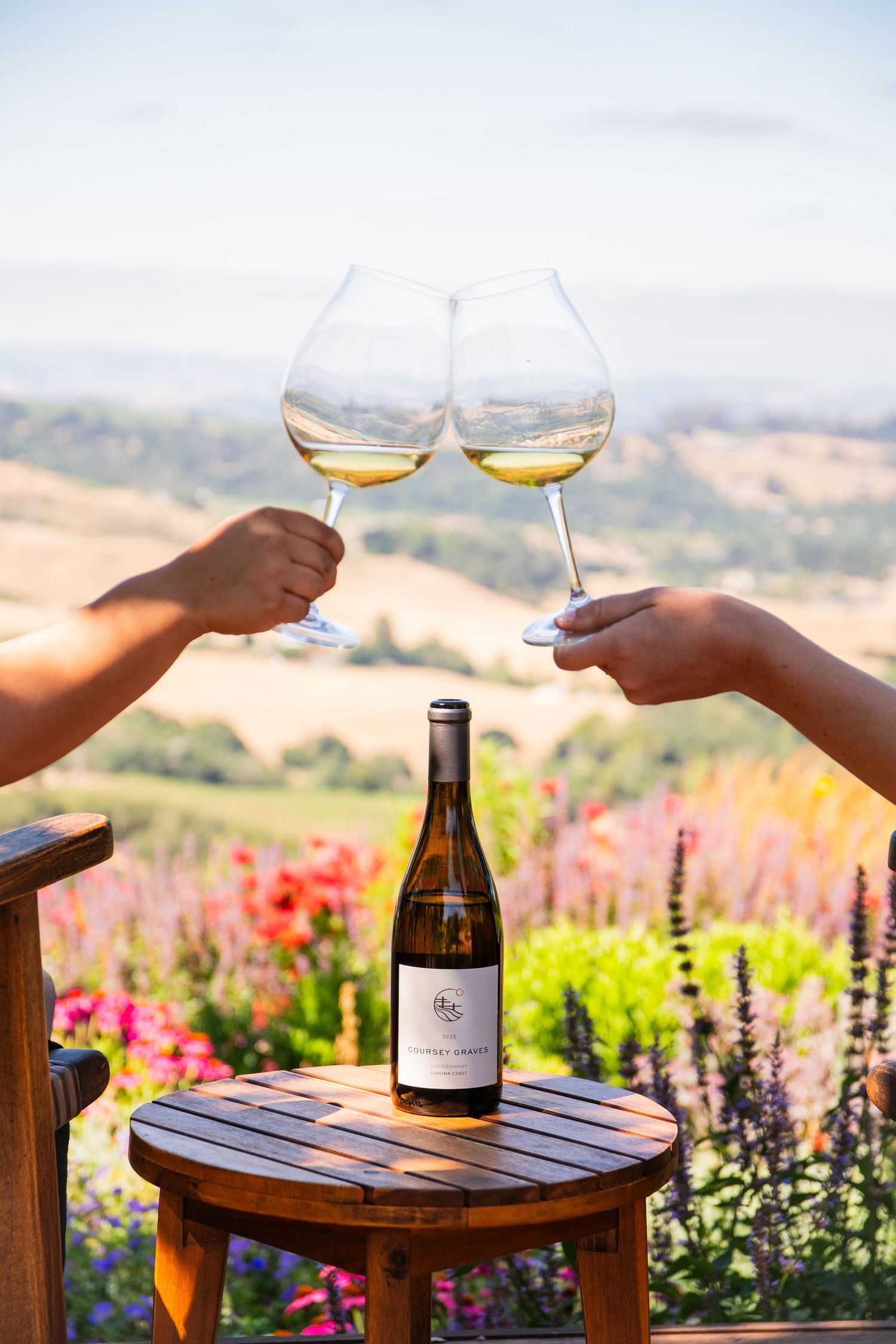Two wine glasses clinking against scenic vineyard backdrop with colorful flowers.