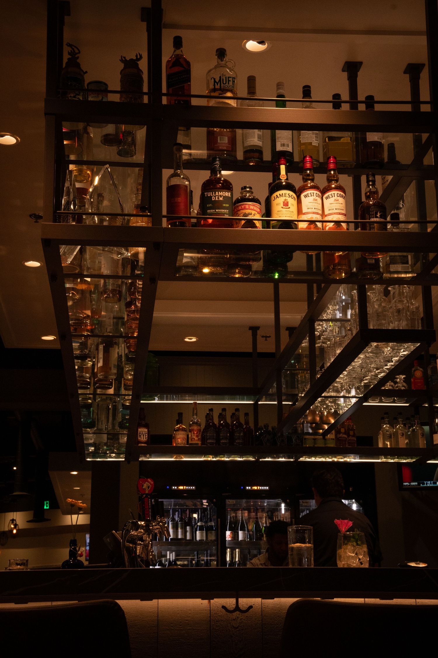 Dimly lit bar interior with multiple shelves displaying bottles of spirits and glassware in an upscale setting.