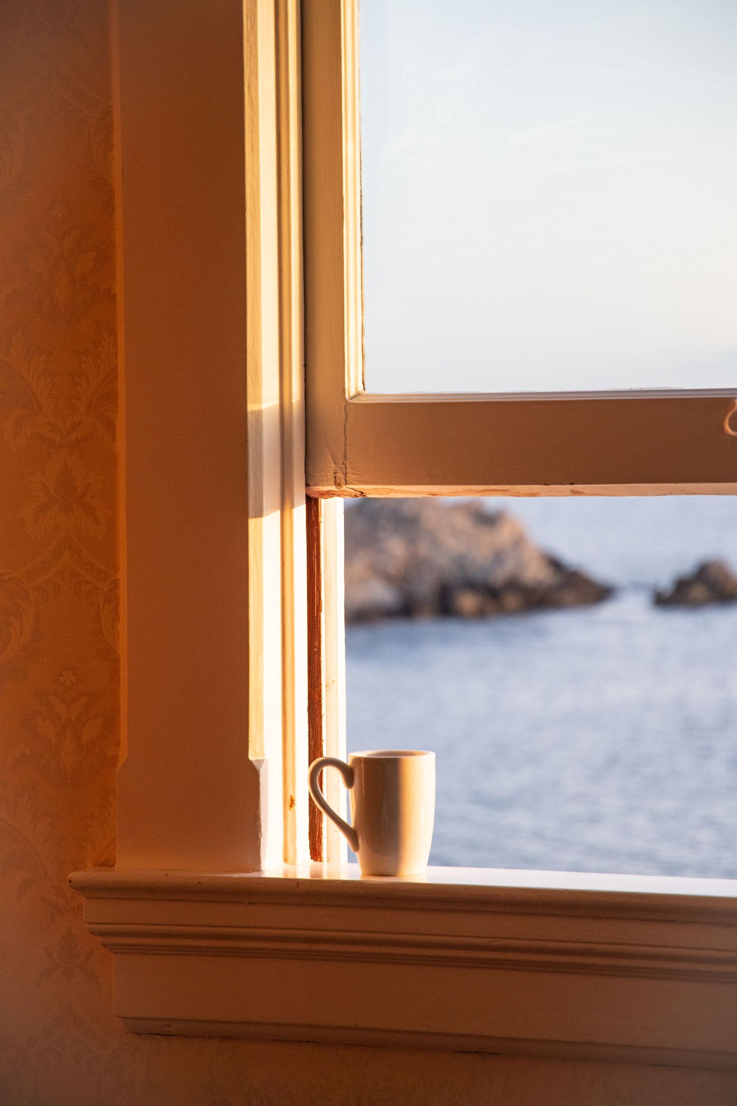 Coffee mug on windowsill overlooking rocky ocean coastline.