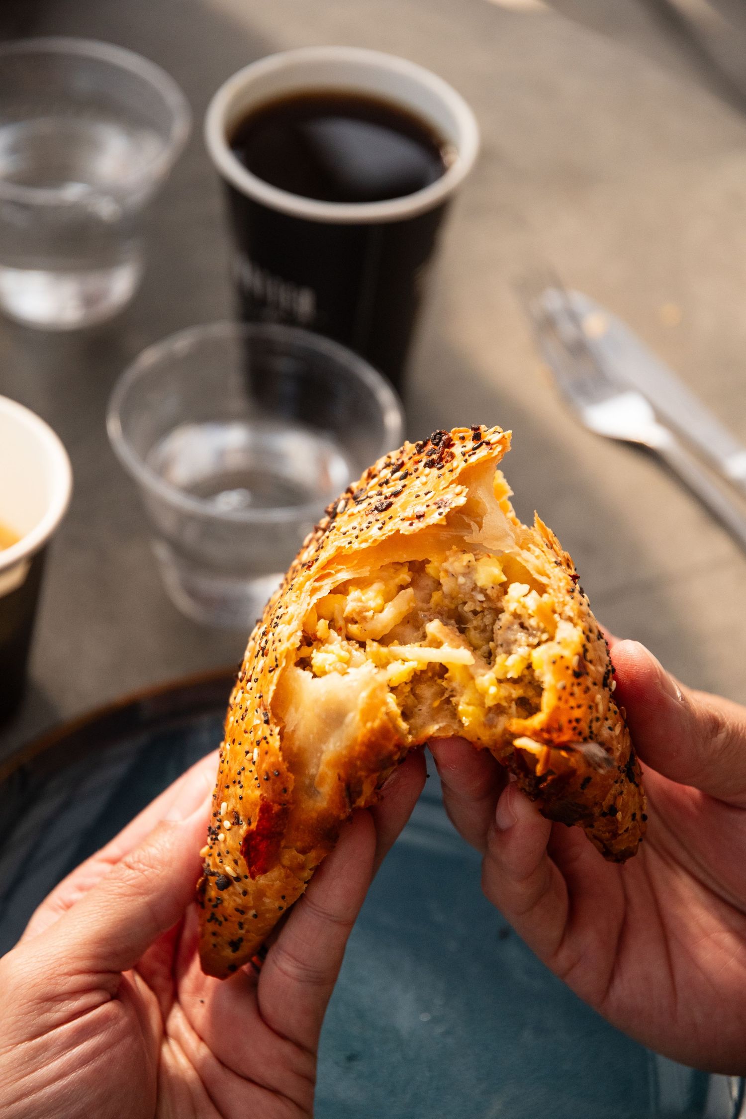Hands breaking open a fresh baked pastry with flaky layers beside coffee cups.