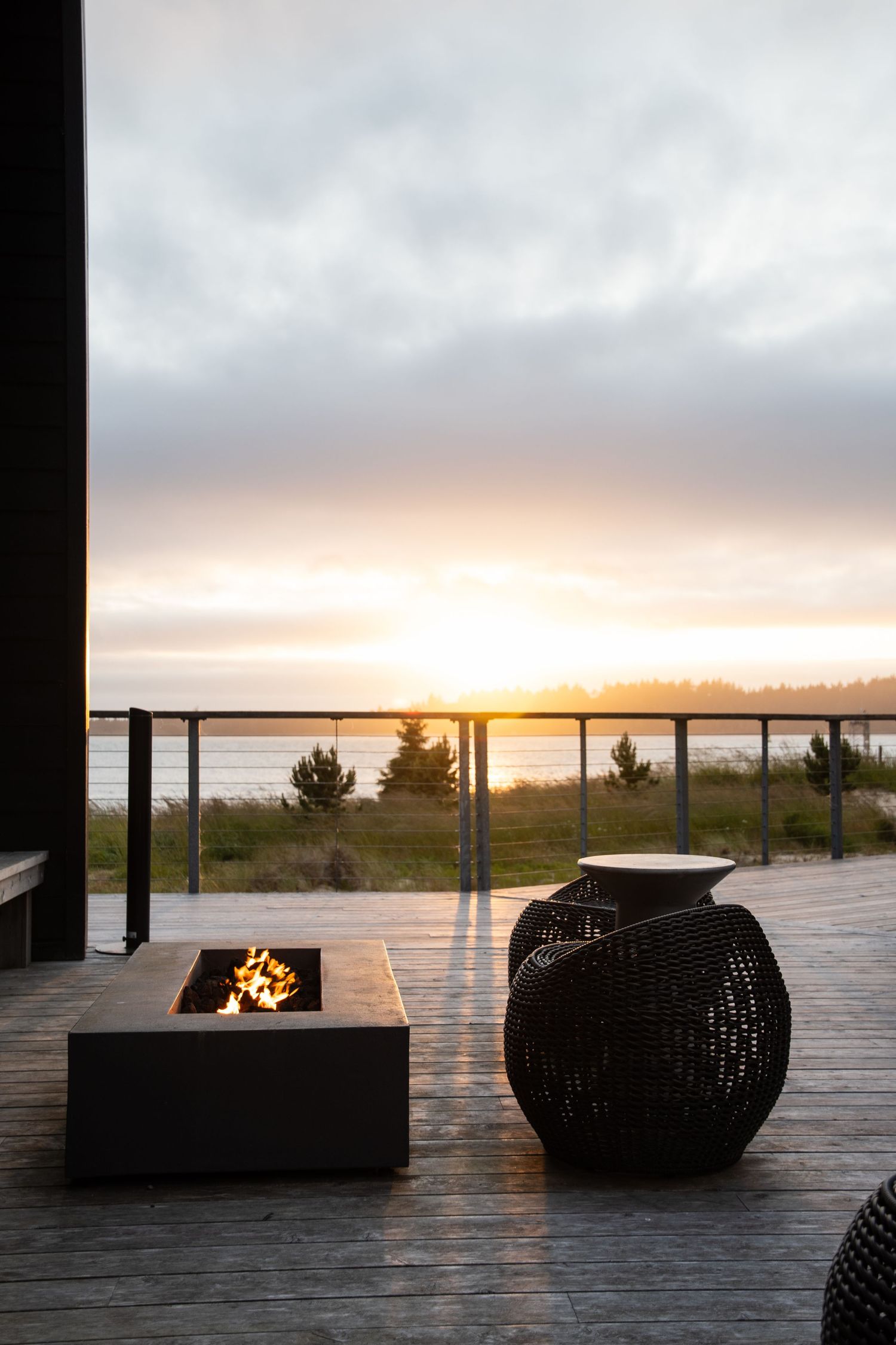 Outdoor deck with modern fire pits and seating silhouetted against sunset ocean view.