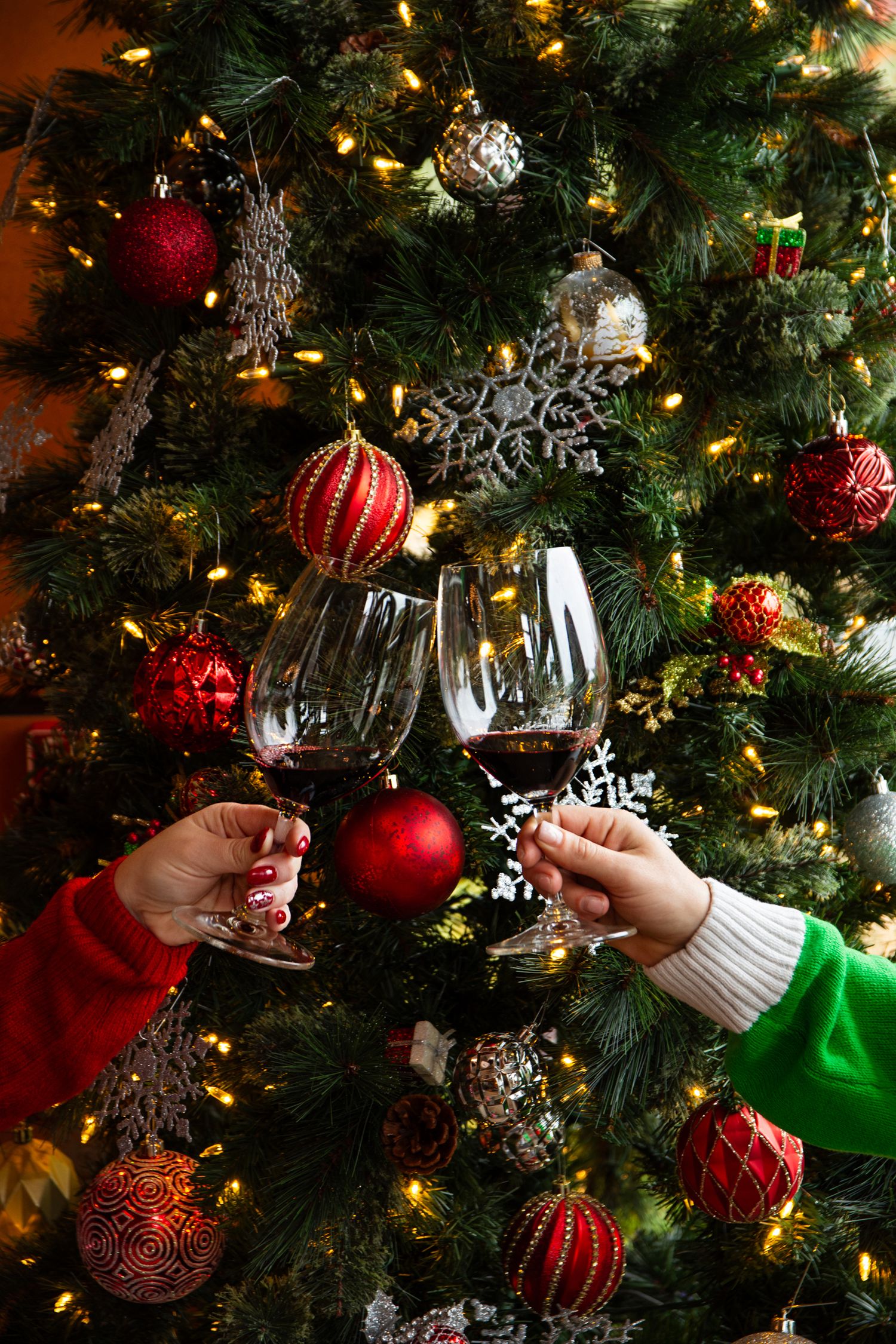 Hands toasting with red wine glasses in front of decorated Christmas tree with ornaments.