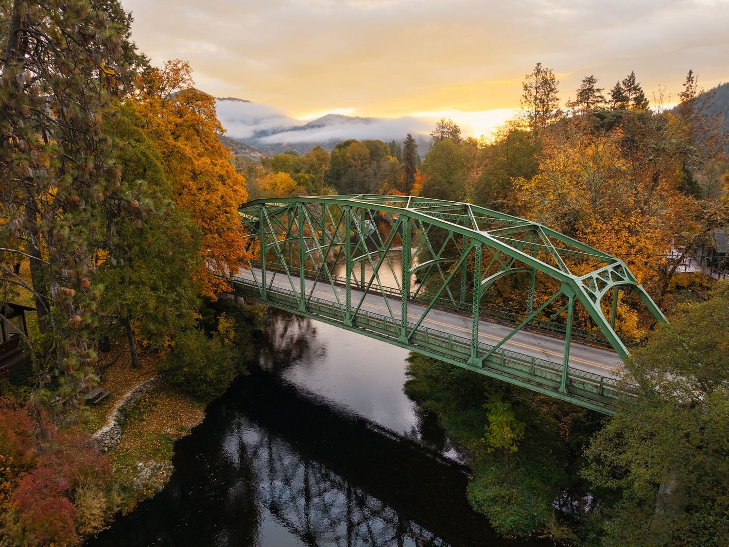 Green steel bridge over a calm river surrounded by autumn foliage with misty mountains at sunset.