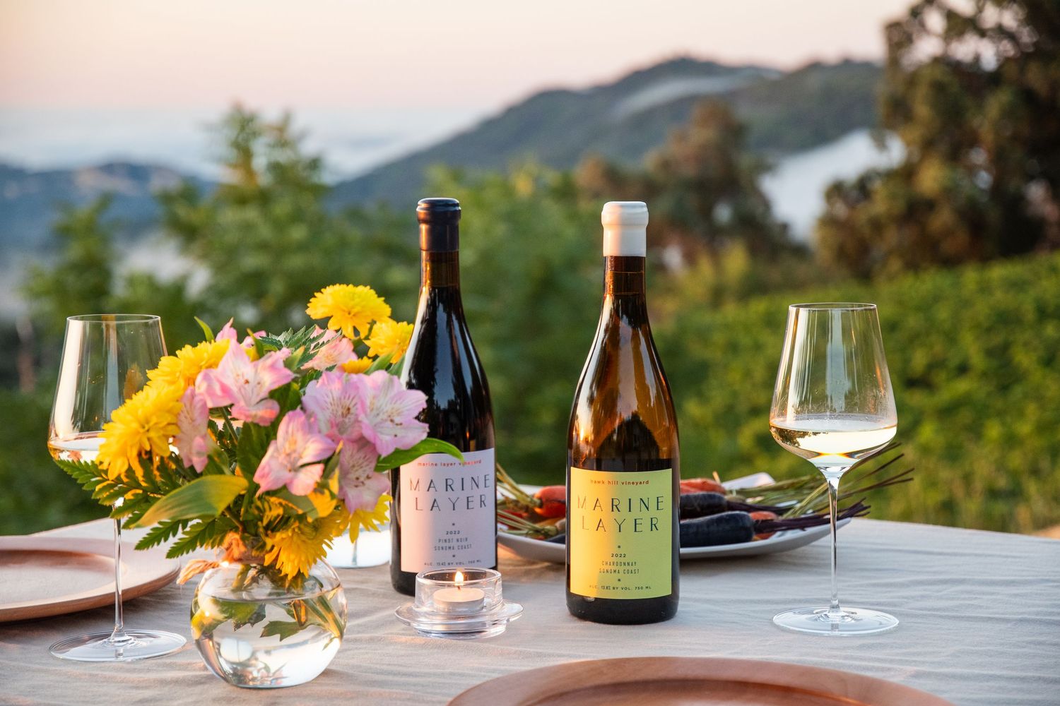 Wine bottles and glasses arranged on an outdoor table with mountain views at sunset.