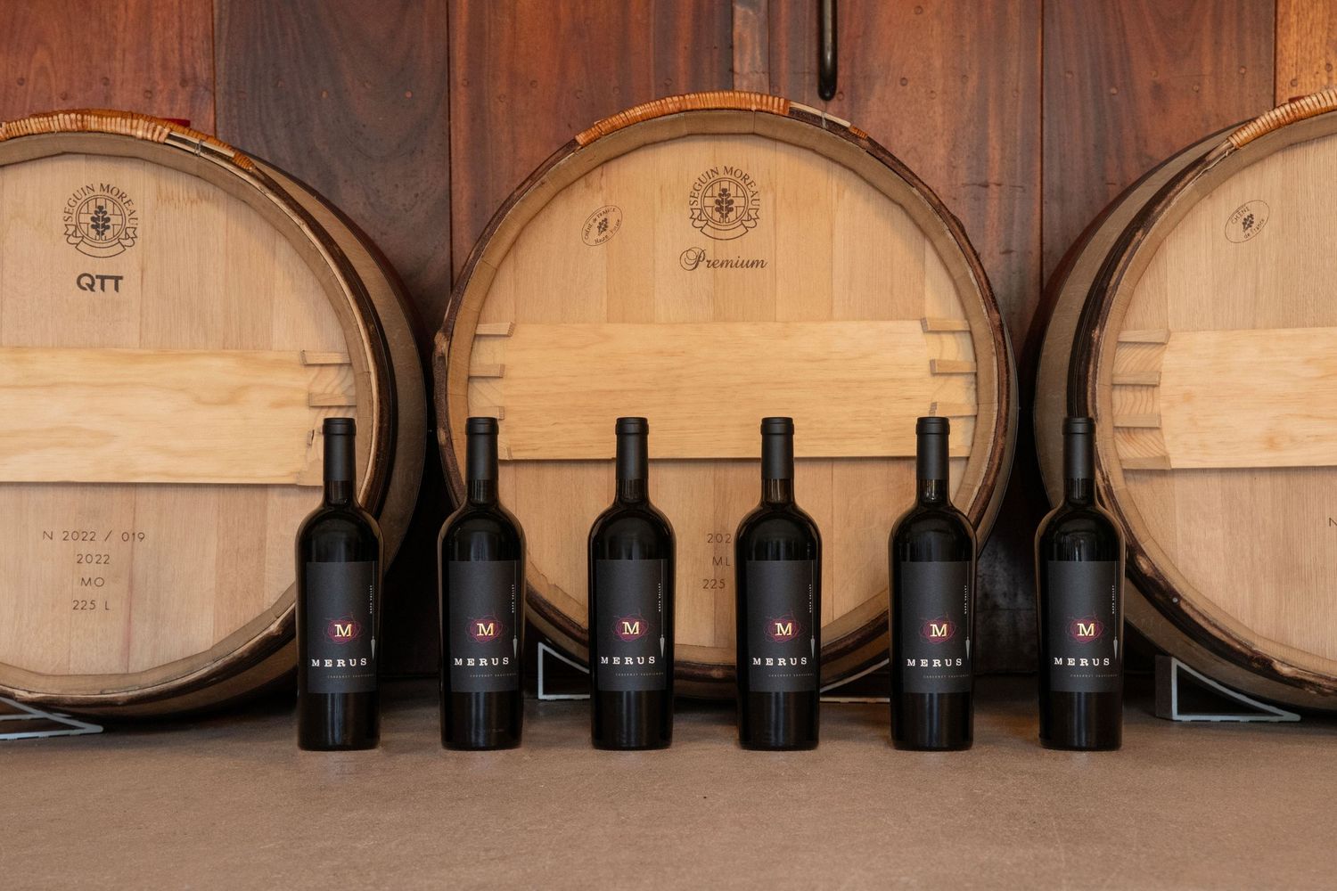 Six dark wine bottles labeled Nodus displayed in front of oak aging barrels in a winery cellar.