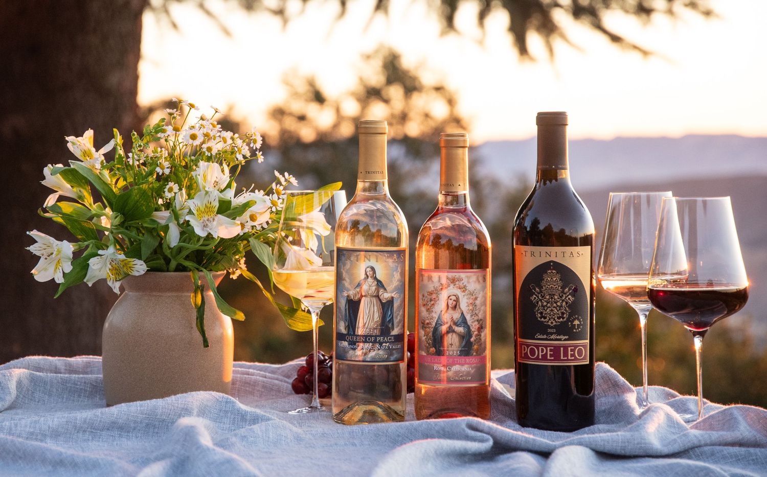 Three wine bottles with ornate labels displayed on a table with white flowers and two glasses of red wine at sunset.