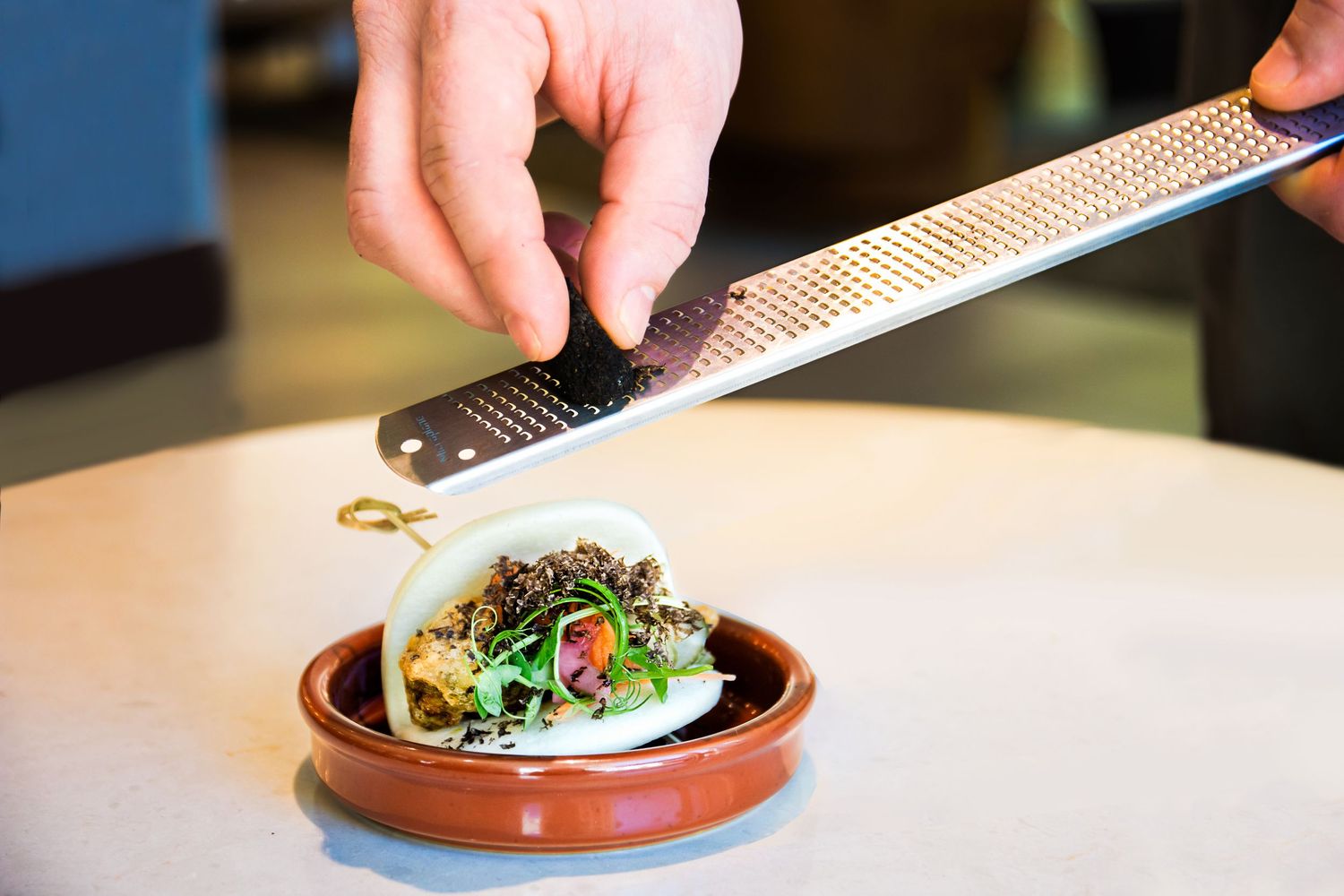 Chef grating fresh garnish over a plated dish in an earthenware bowl.