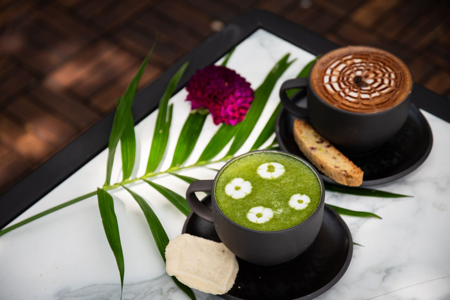 Two artisanal lattes with latte art served with cookies and tropical palm leaf decoration on marble surface.