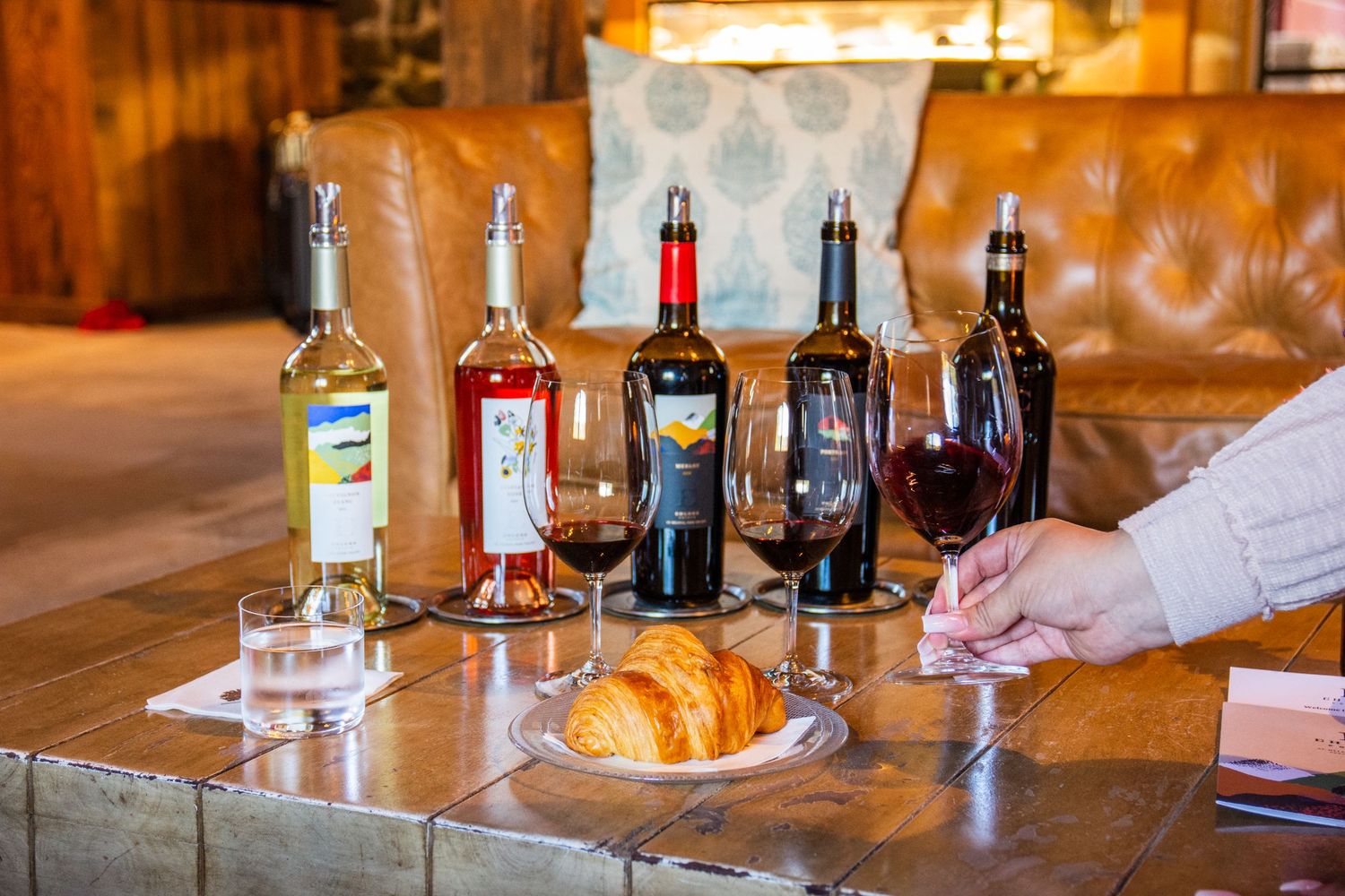Wine tasting setup with four bottles, filled glasses, a croissant, and a hand holding a glass of red wine.
