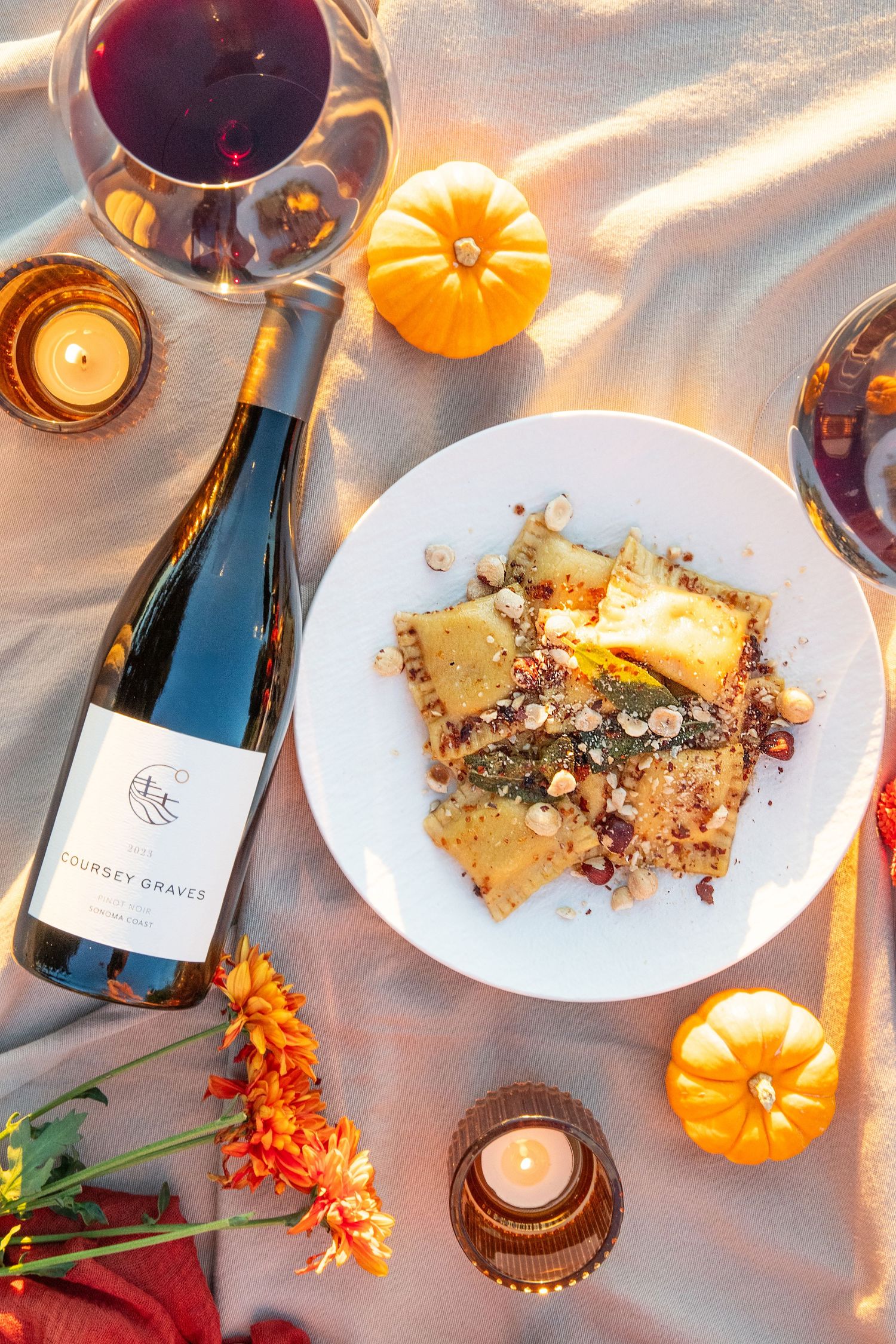 Elegant table setting with wine bottles, mini pumpkins, and plated desserts in warm candlelight.