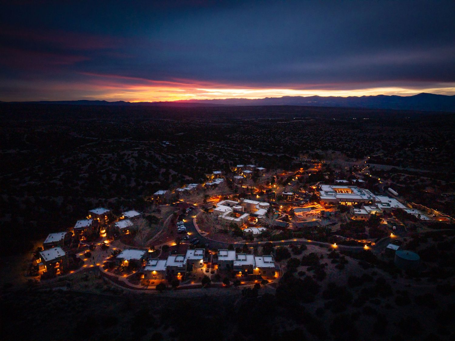 Aerial drone view of a glowing desert town at dusk with vibrant sunset sky and mountain silhouettes in New Mexico.