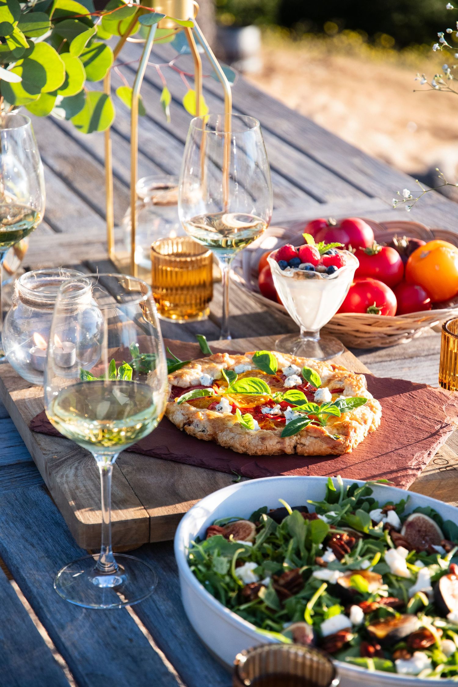 Outdoor dining setup with wine, fresh salad, and flatbread pizza on rustic wooden table.
