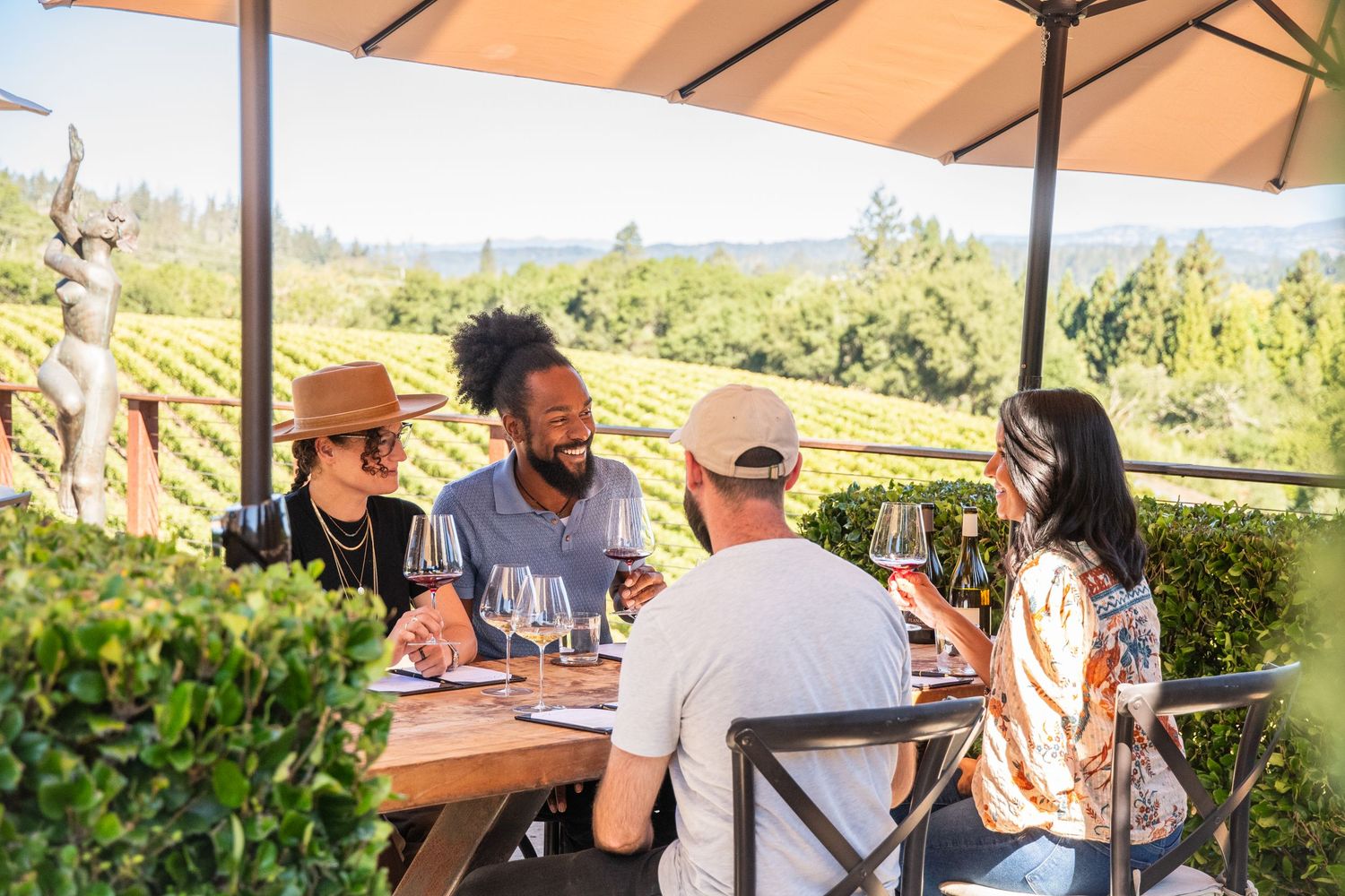 Four friends enjoy wine tasting at an outdoor vineyard patio surrounded by lush green vines on a sunny day.