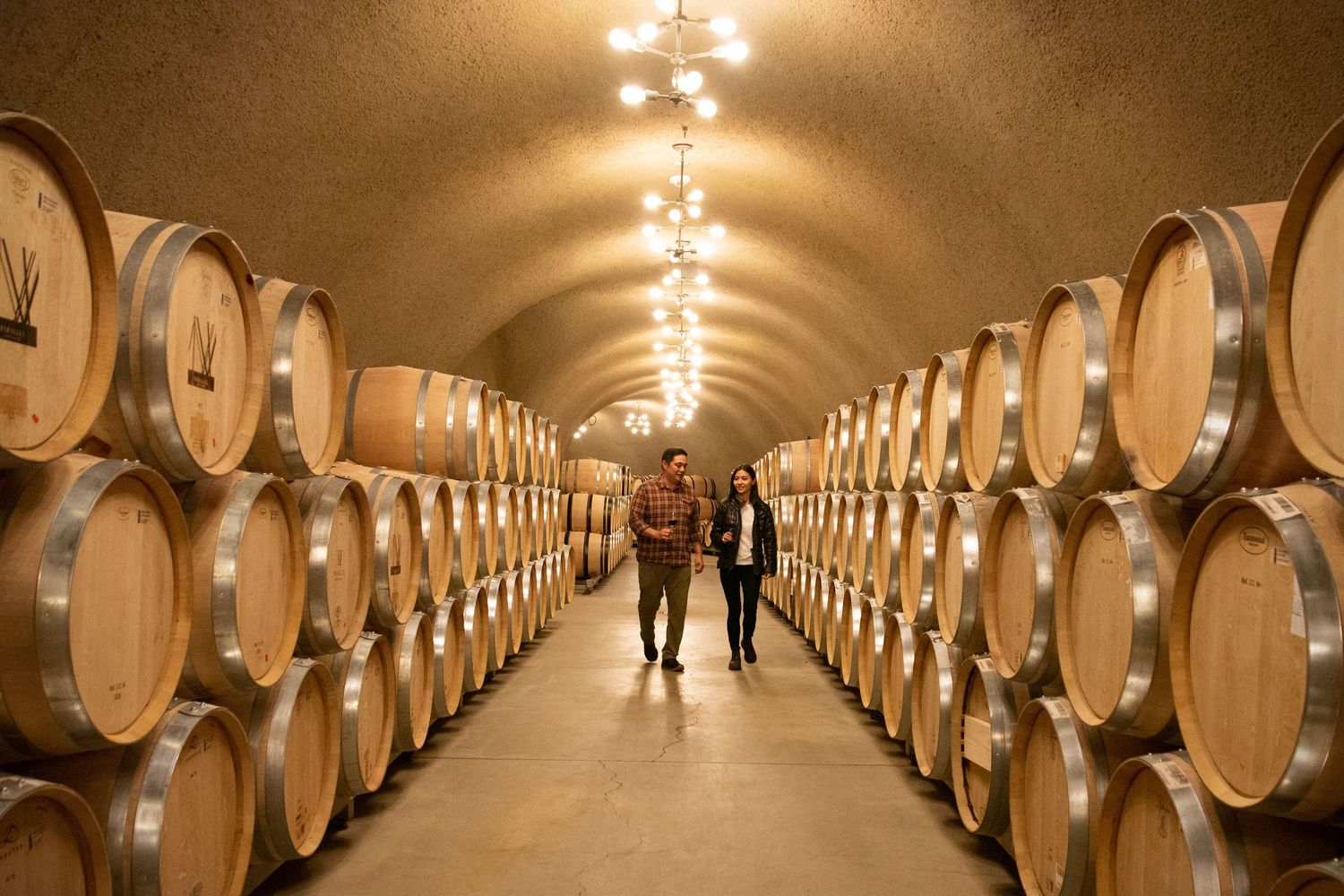 Wine barrels line a dimly lit cellar tunnel with overhead lights creating an atmospheric perspective.