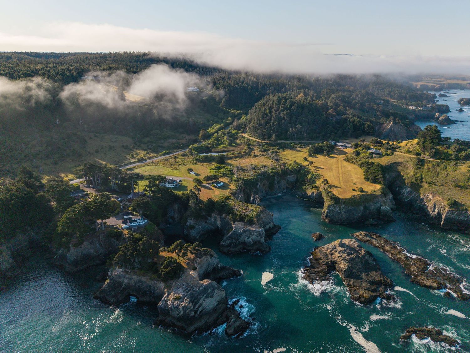 Aerial view of a rugged coastal landscape with rocky cliffs, turquoise waters, and lush green hills under a cloudy sky.