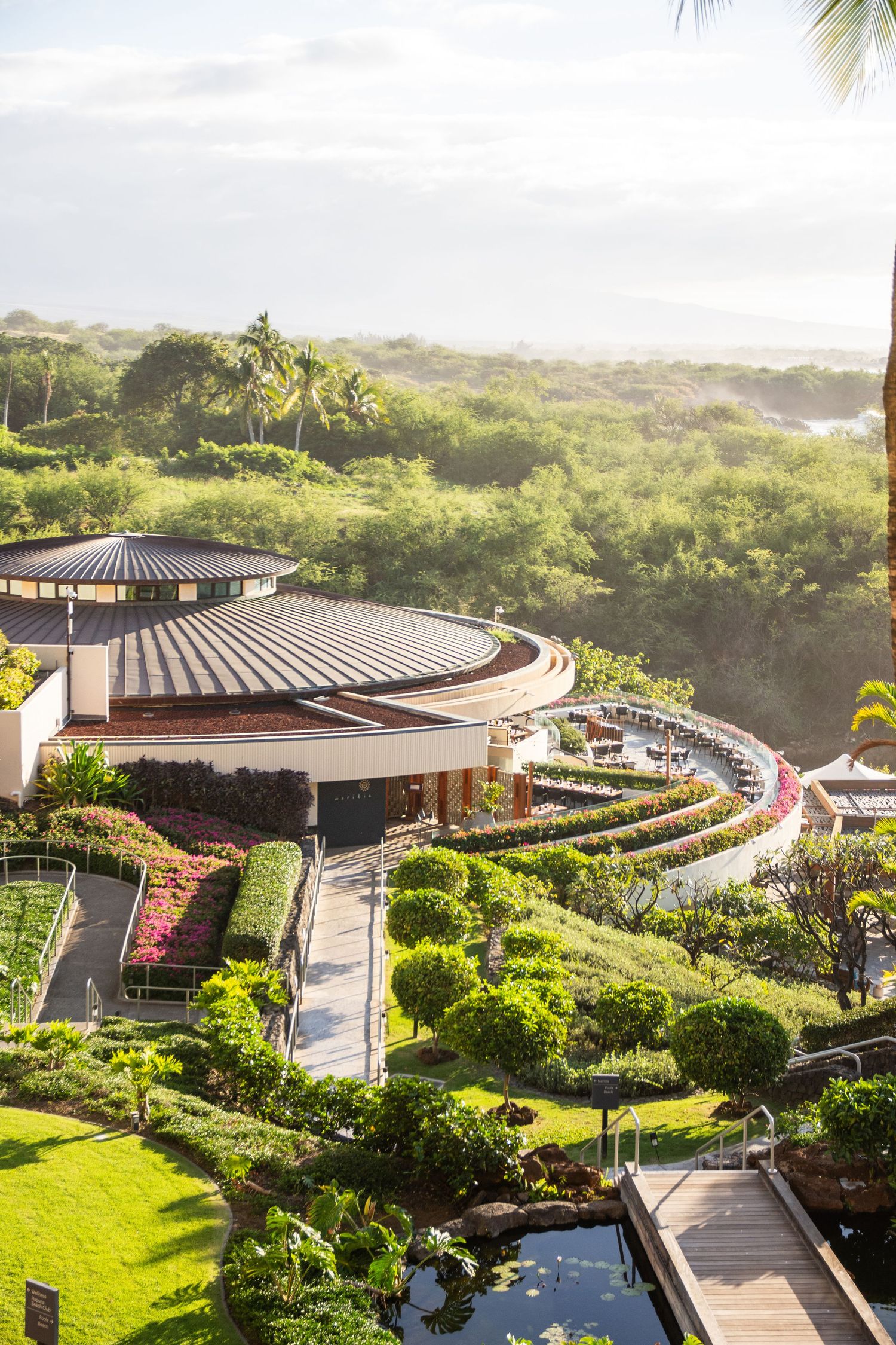 Aerial view of a circular resort building surrounded by lush tropical gardens and palm trees near the ocean coastline.