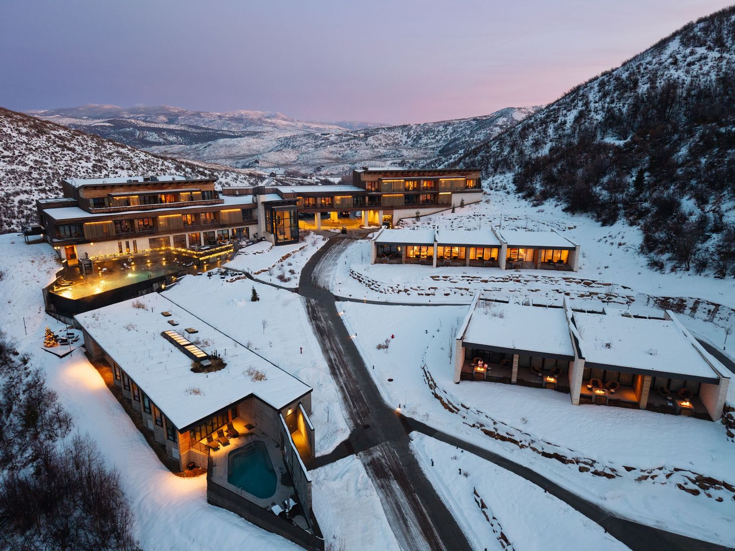 Aerial view of a luxury mountain resort blanketed in snow at dusk, with warm glowing lights and snowy hills.