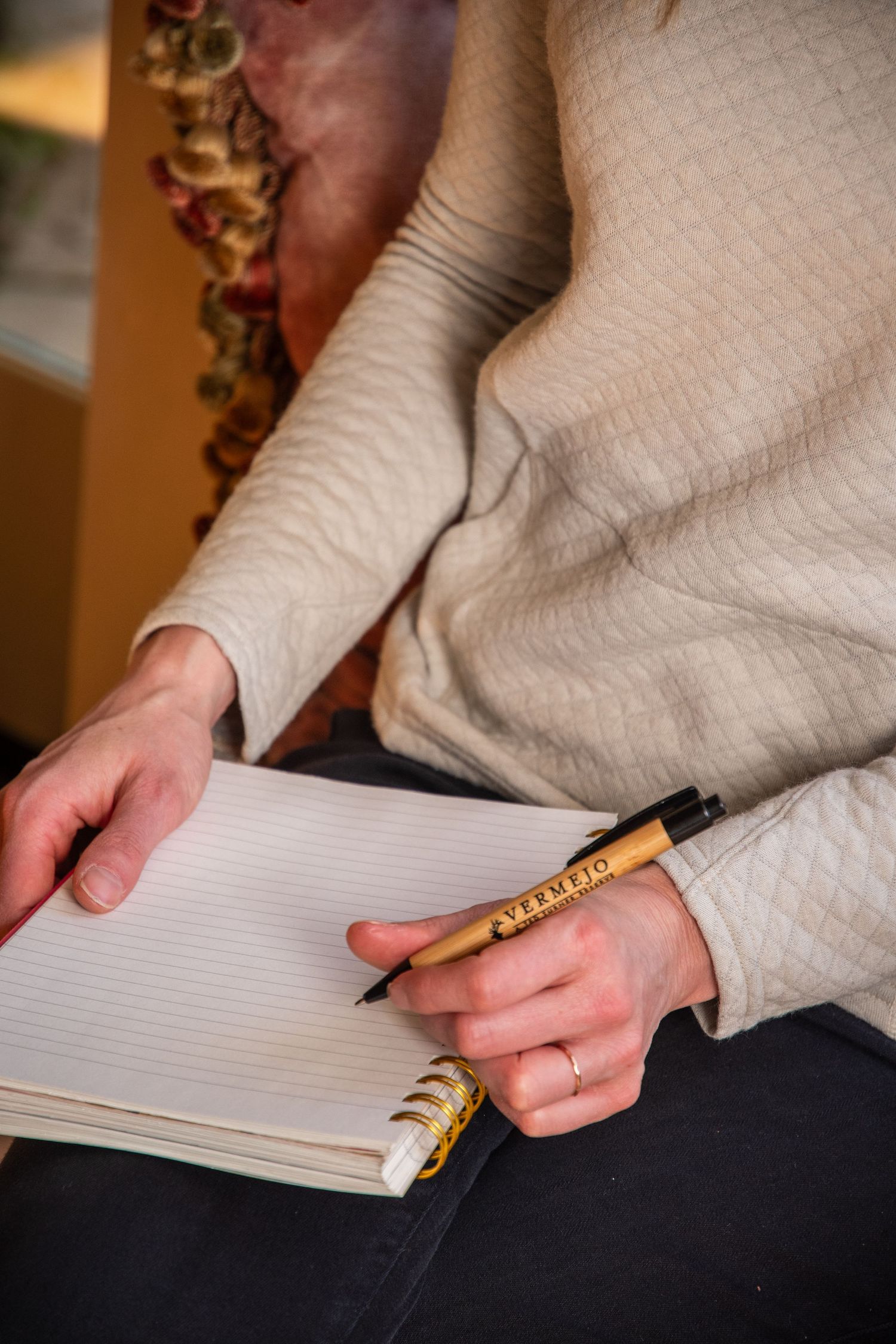 Person in white sweater writing in a spiral notebook with a decorative wooden pen, wearing a ring.