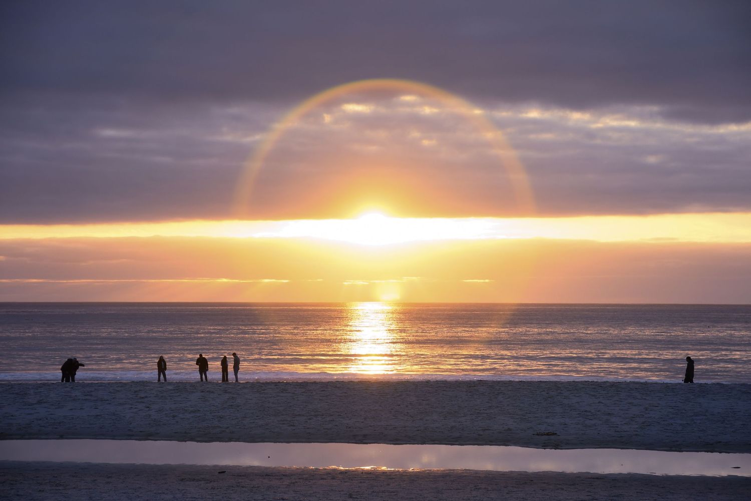 Golden sunset over beach with rainbow arc and silhouettes on shore.