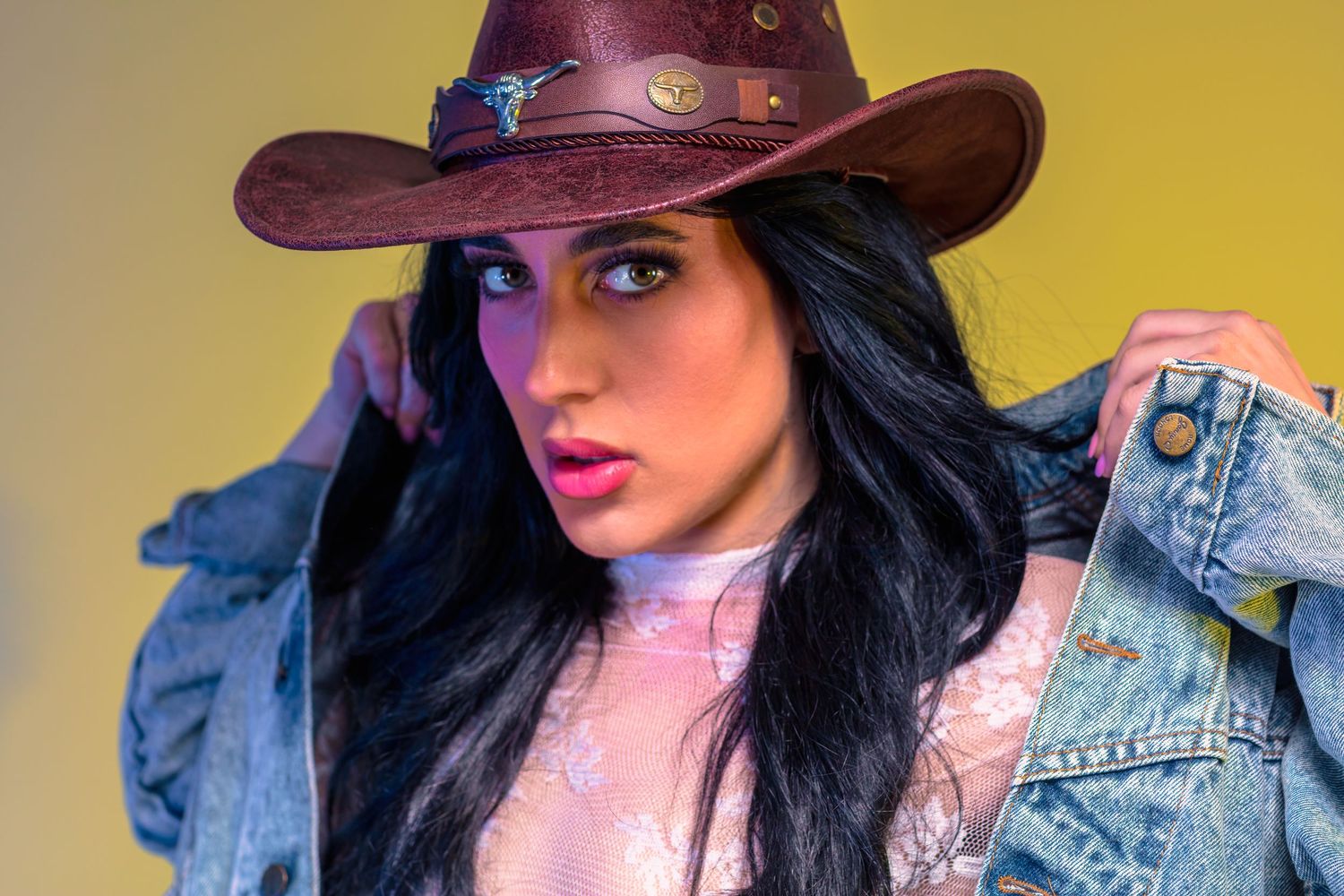 Young woman in brown cowboy hat and denim jacket posing against yellow background.