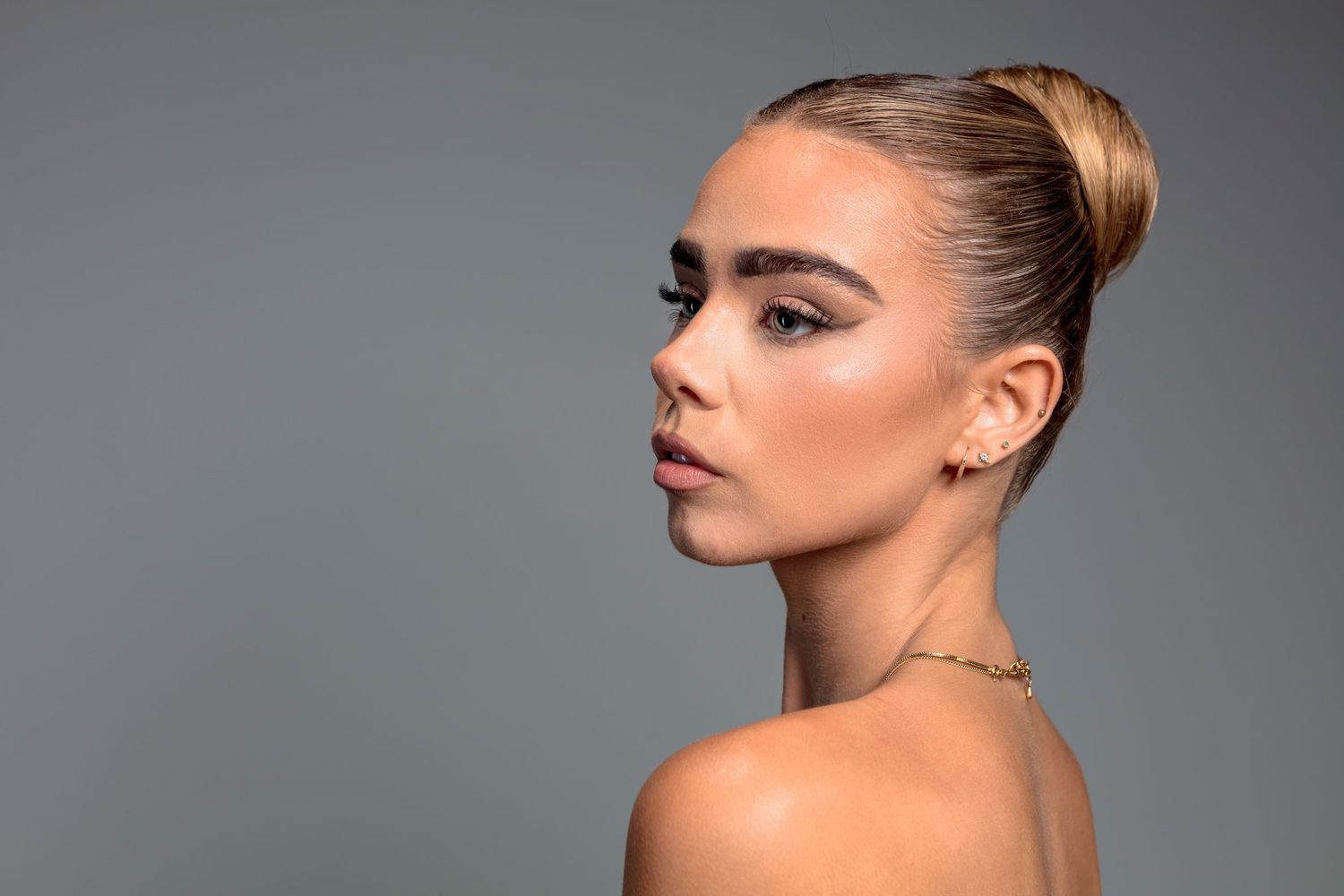 Young woman with sleek bun hairstyle, bold brows, gold earring, and bare shoulders poses against gray background.