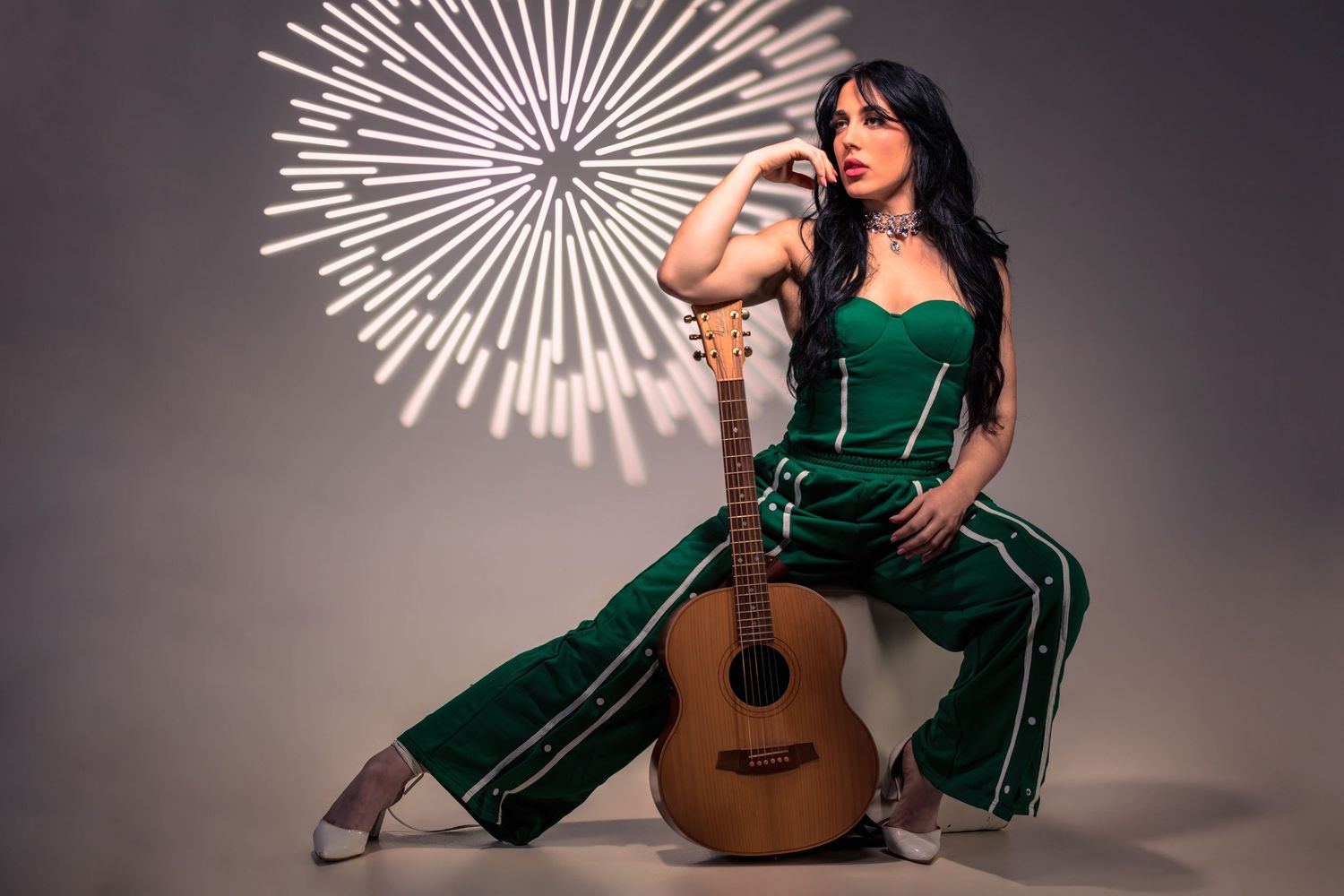 Woman in green jumpsuit posing with acoustic guitar against gray background with starburst light design.
