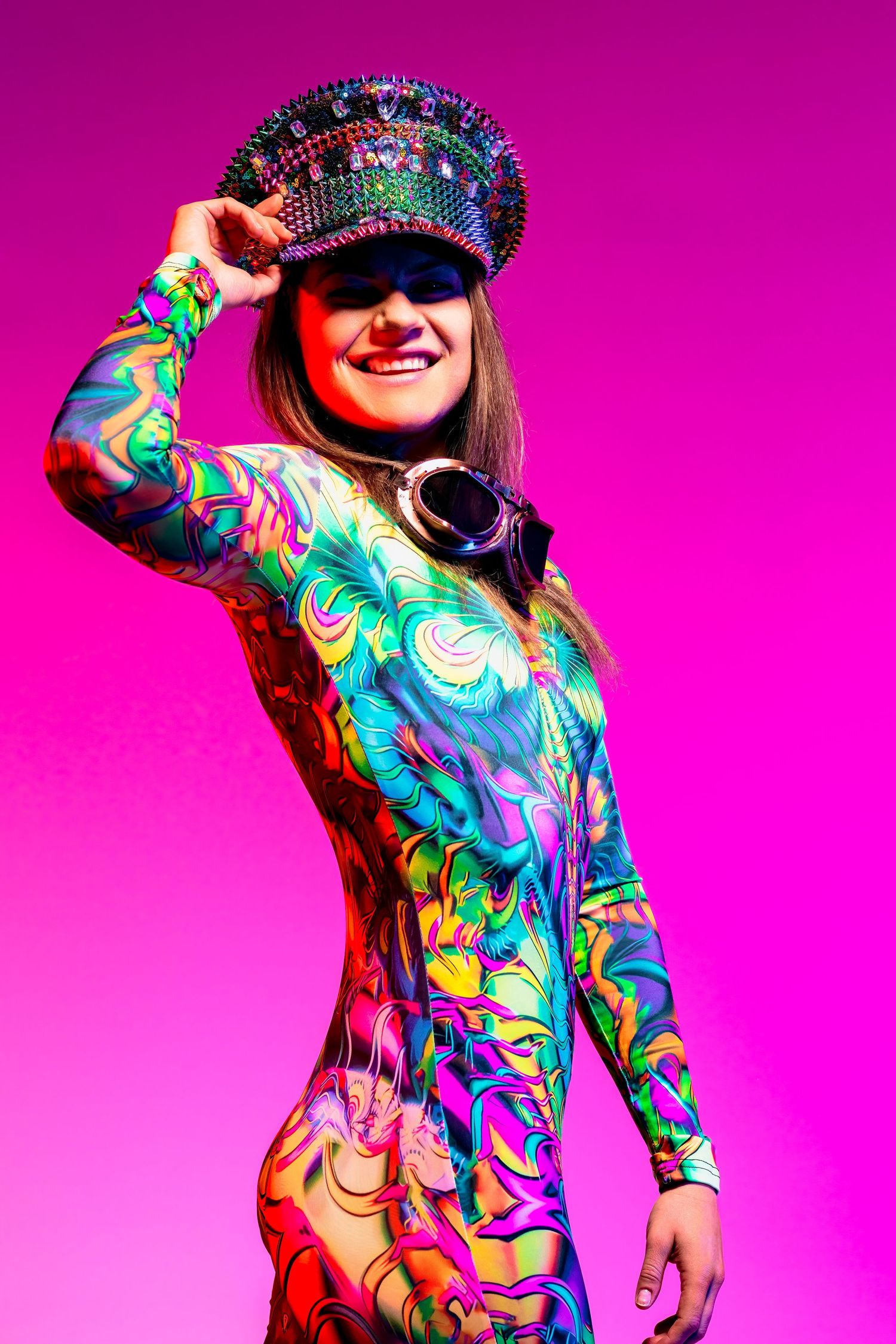 Person wearing colorful psychedelic bodysuit and embellished cap poses against vibrant pink background.