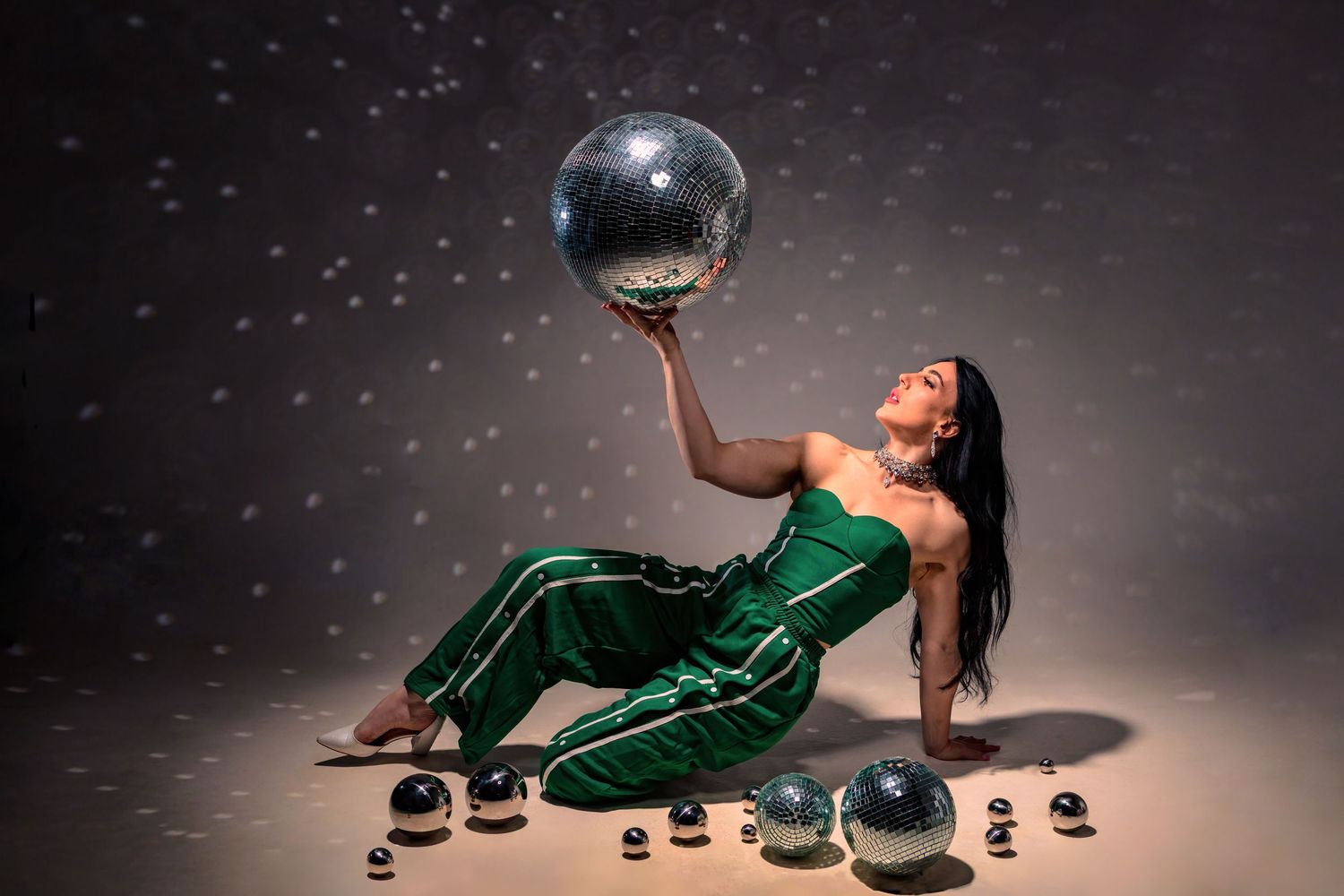 Woman in green jumpsuit poses on floor holding large disco ball aloft, surrounded by smaller mirror balls.