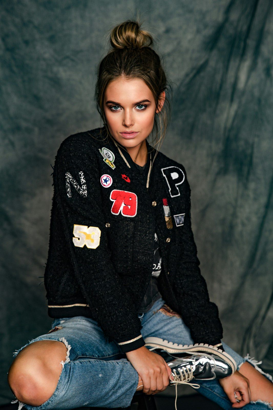 Model wearing varsity jacket with patches and ripped jeans in moody studio portrait against dark backdrop.