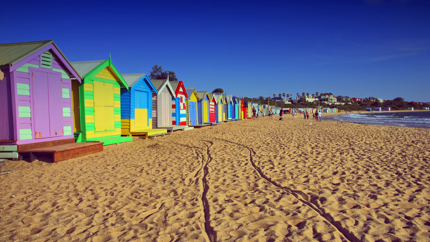 Colorful beach huts line the sandy shore of Brighton beach under bright blue sky.