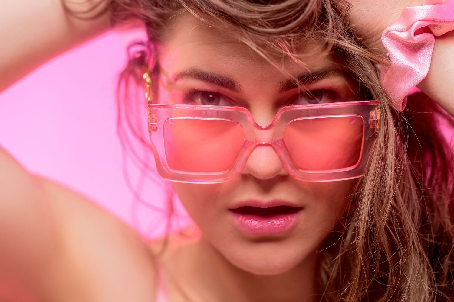 Young woman wearing oversized pink tinted square sunglasses poses against a vibrant pink background.