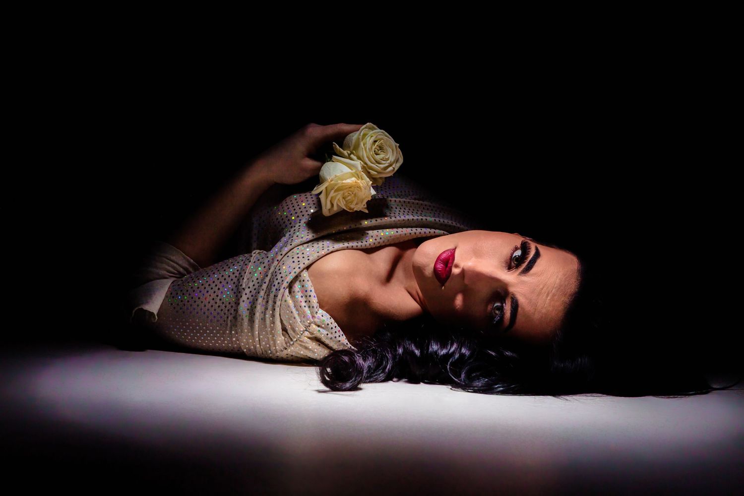 Woman in silver dress lying on floor holding cream roses with dramatic dark background lighting.