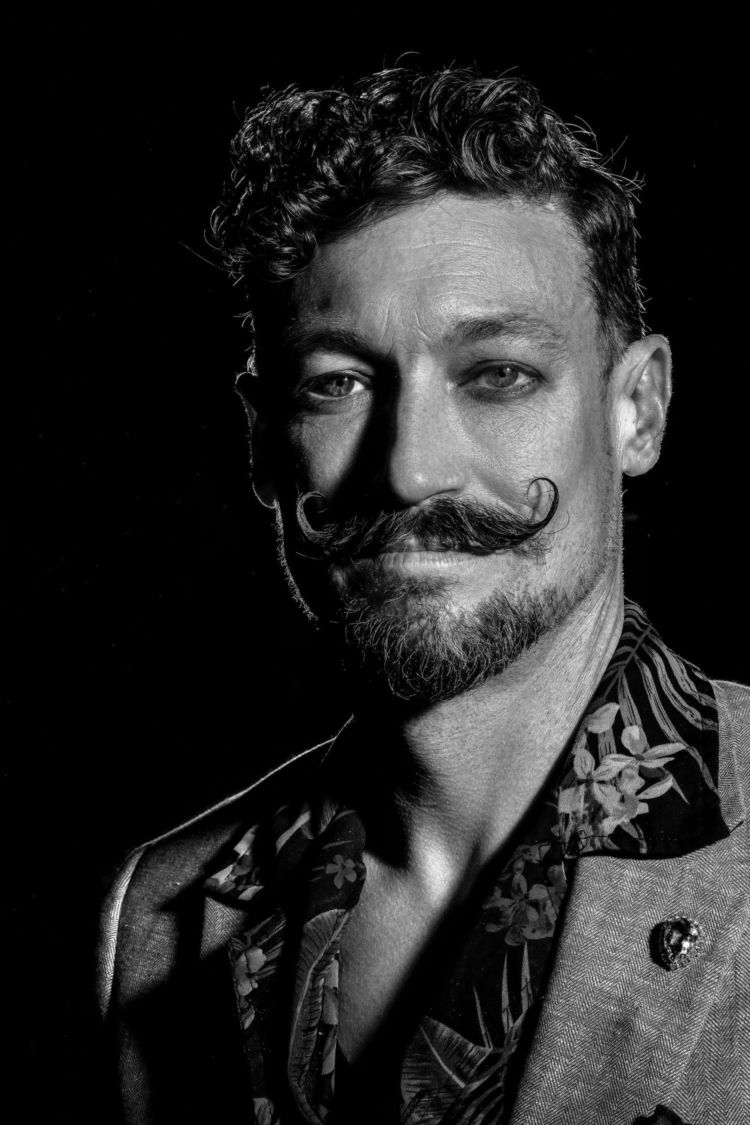 Black and white portrait of a man with a curled mustache, beard, and curly hair wearing a floral shirt.