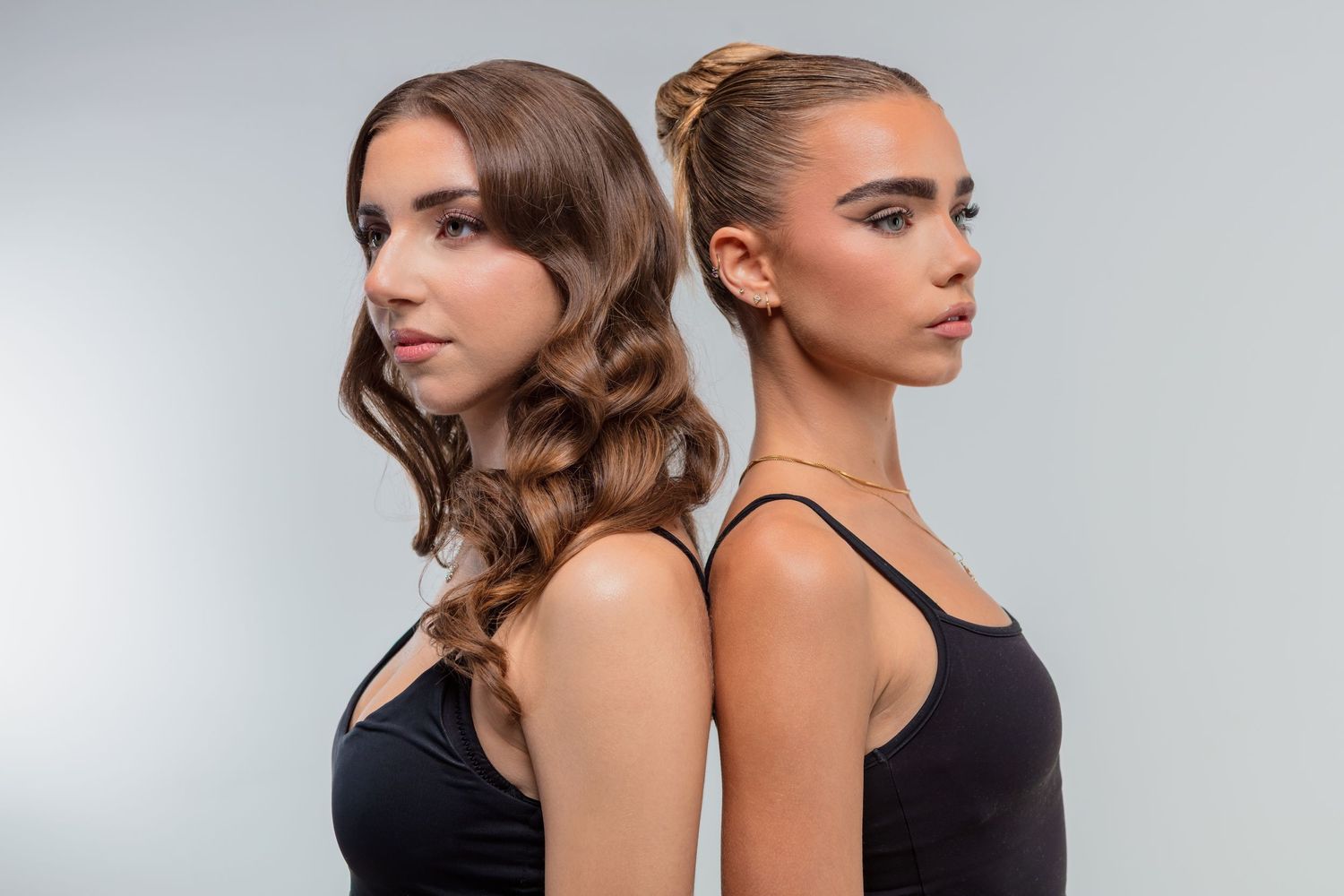 Two young women in black tops stand back-to-back, one with curly hair and one with an updo, against a gray background.
