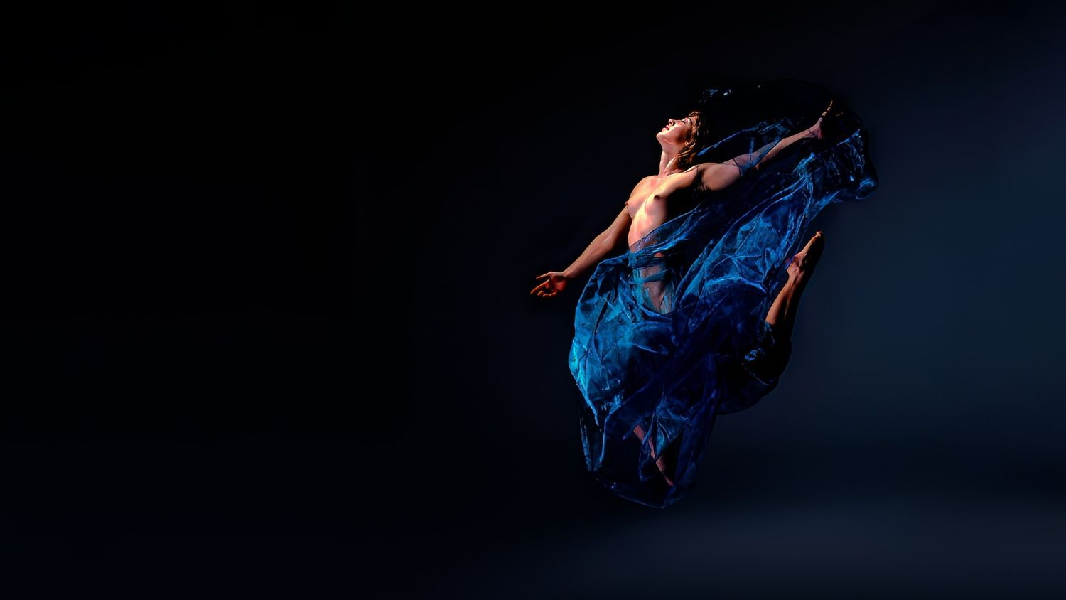 A dancer in flowing blue fabric performs an elegant aerial movement against a dark background.