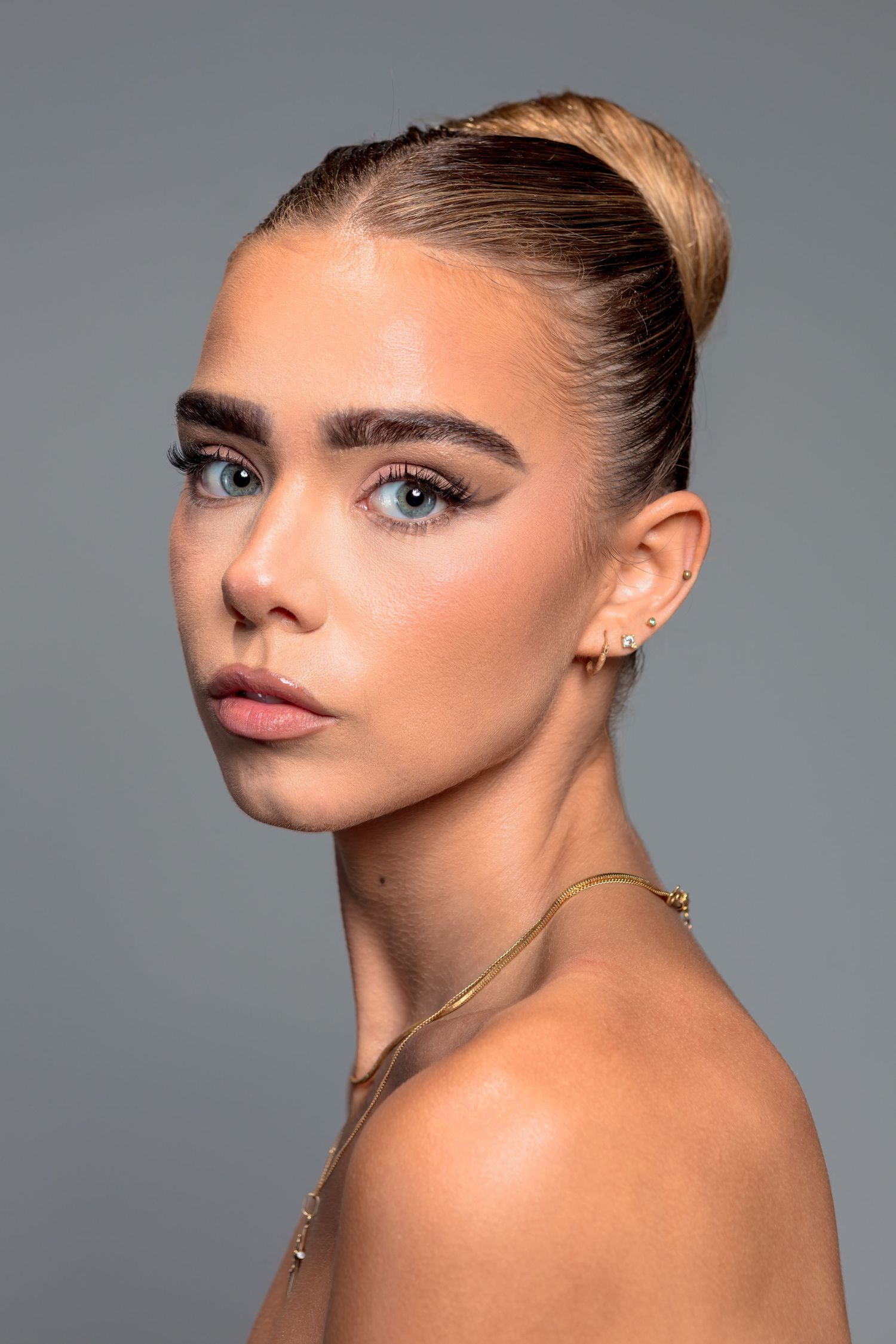 Young woman with blue eyes, sleek bun hairstyle, gold jewelry, and natural glam makeup poses against gray background.