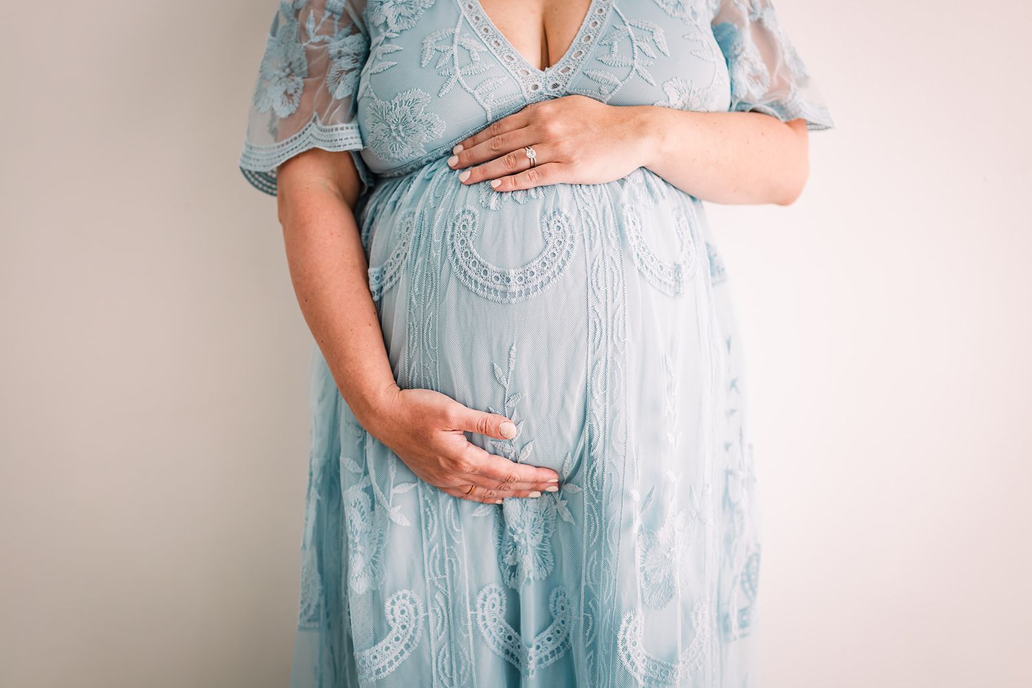 Pregnant woman wearing a light blue lace maternity dress cradles her baby bump against neutral background in a studio in Bedford TX