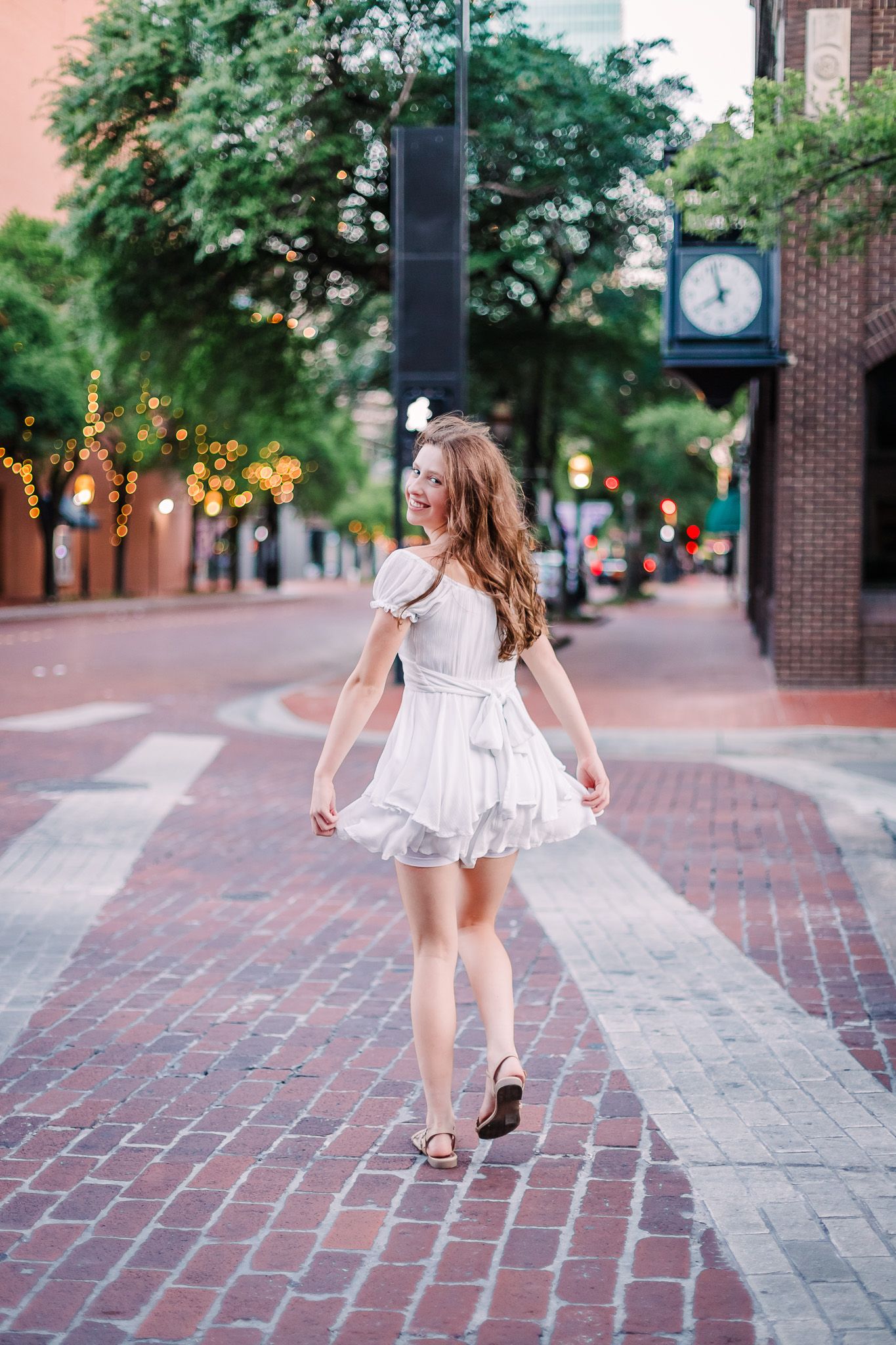 A child in a white dress skips along a brick paved walkway lined with trees and buildings on a summer day at sundance square in fort worth
