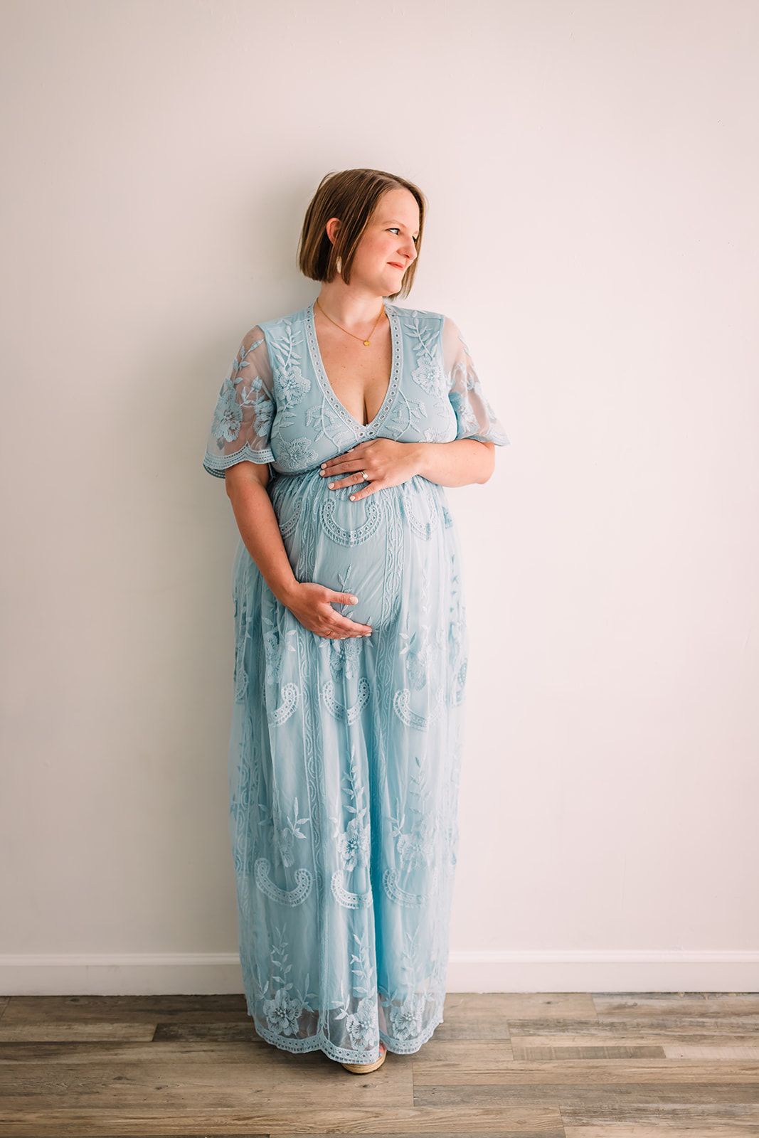 A pregnant woman in a light blue lace maternity gown stands against a neutral wall with hardwood floors.
