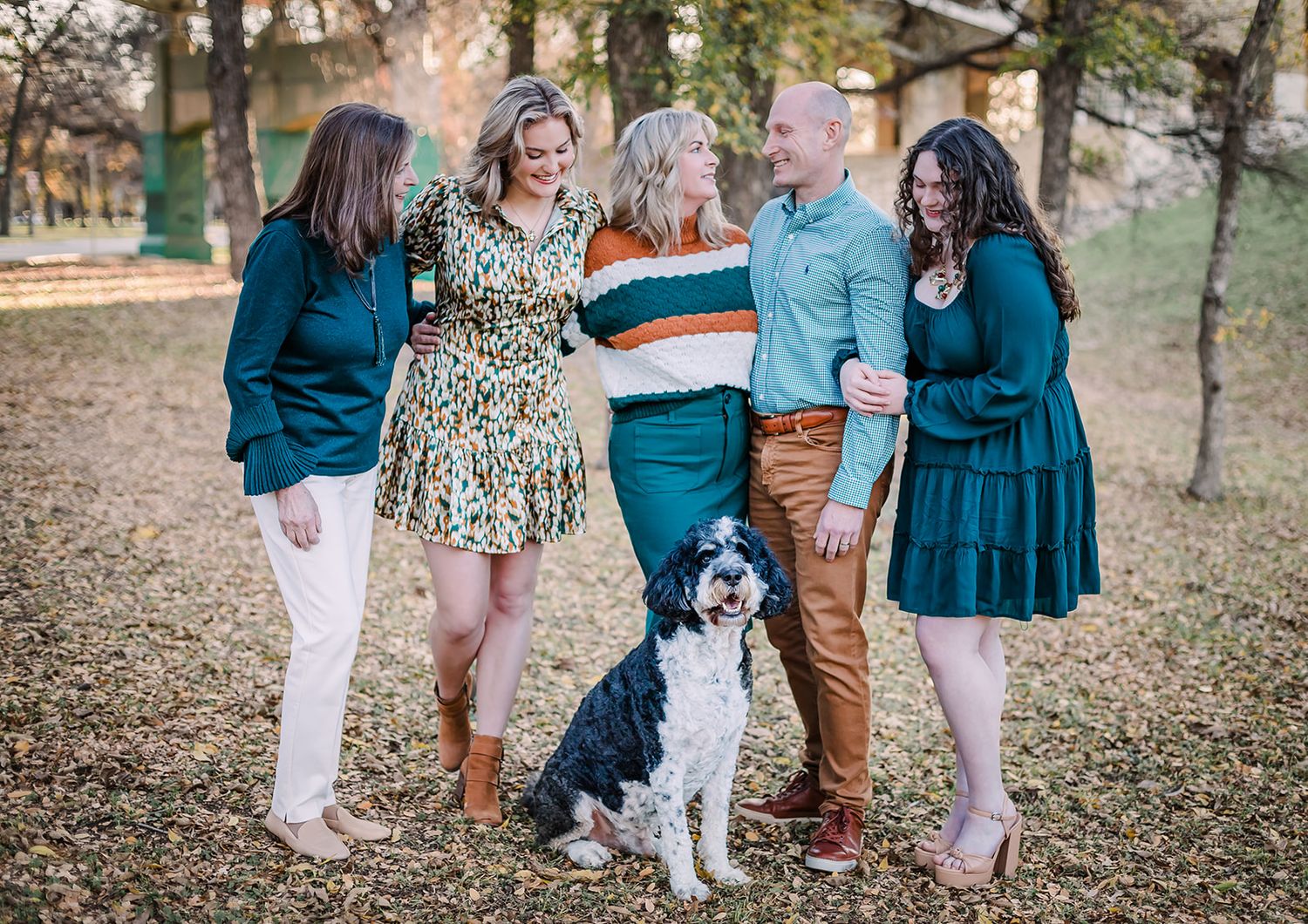 Group in teal and patterned outfits posing with their black and white dog in a wooded setting.