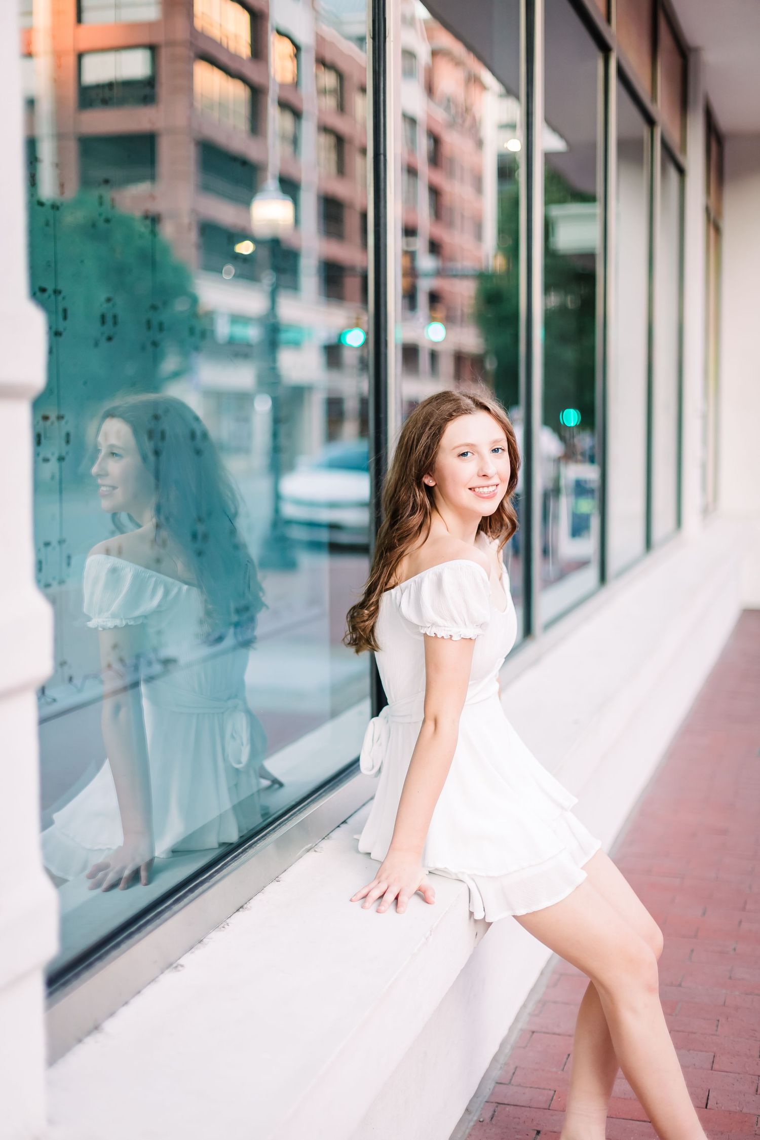 senior girl in white dress poses elegantly against modern glass storefront in Sundance Square Fort Worth TX