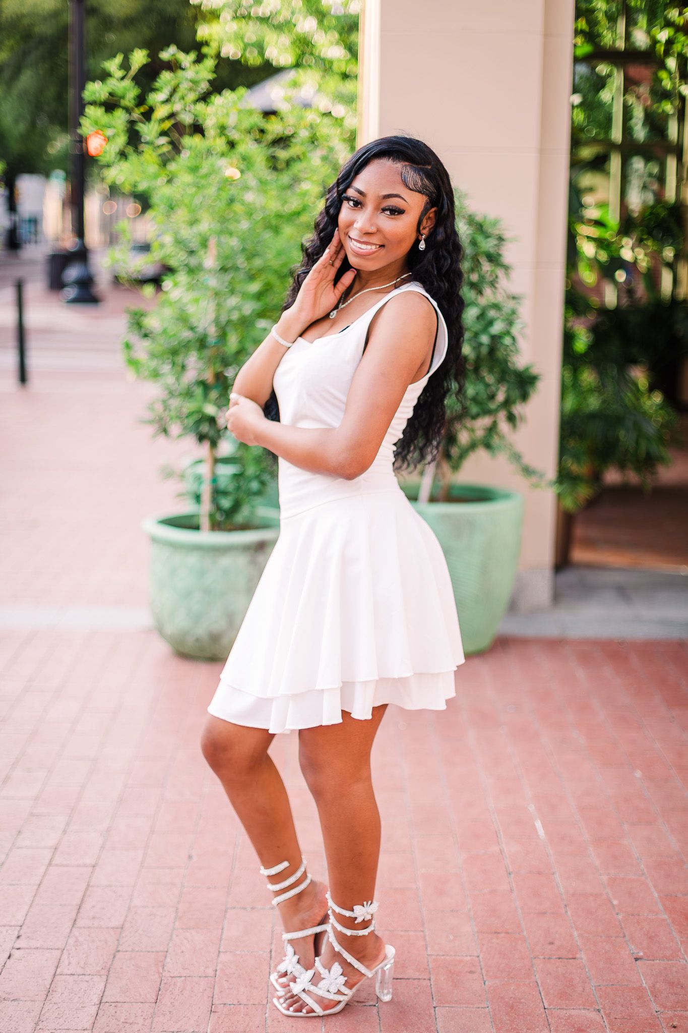A person poses outdoors in a flowy white summer dress and strappy sandals against a pink wall and greenery.