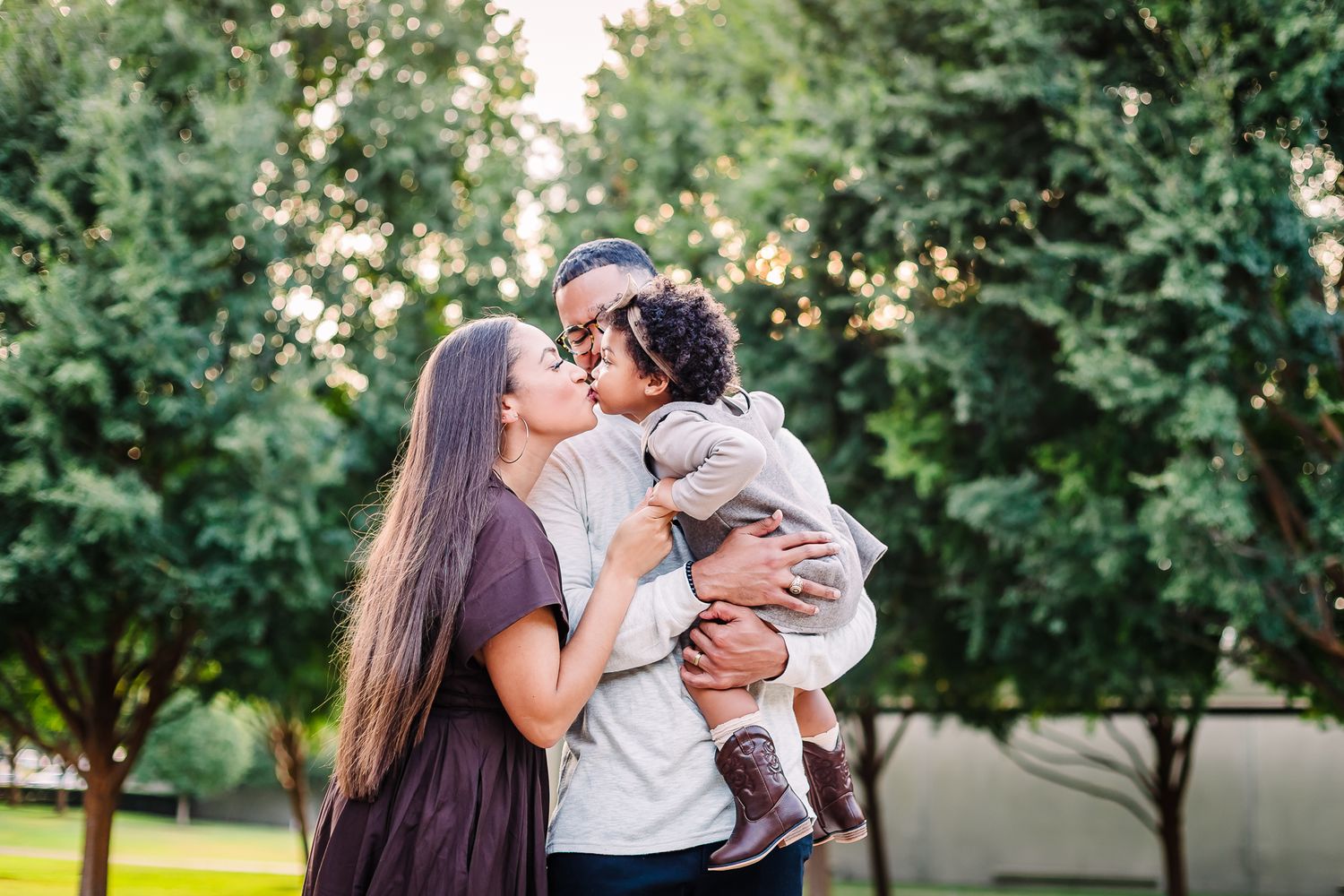 Loving family poses together in front of green trees during outdoor photo session at sunset at Kimbell Art Studio