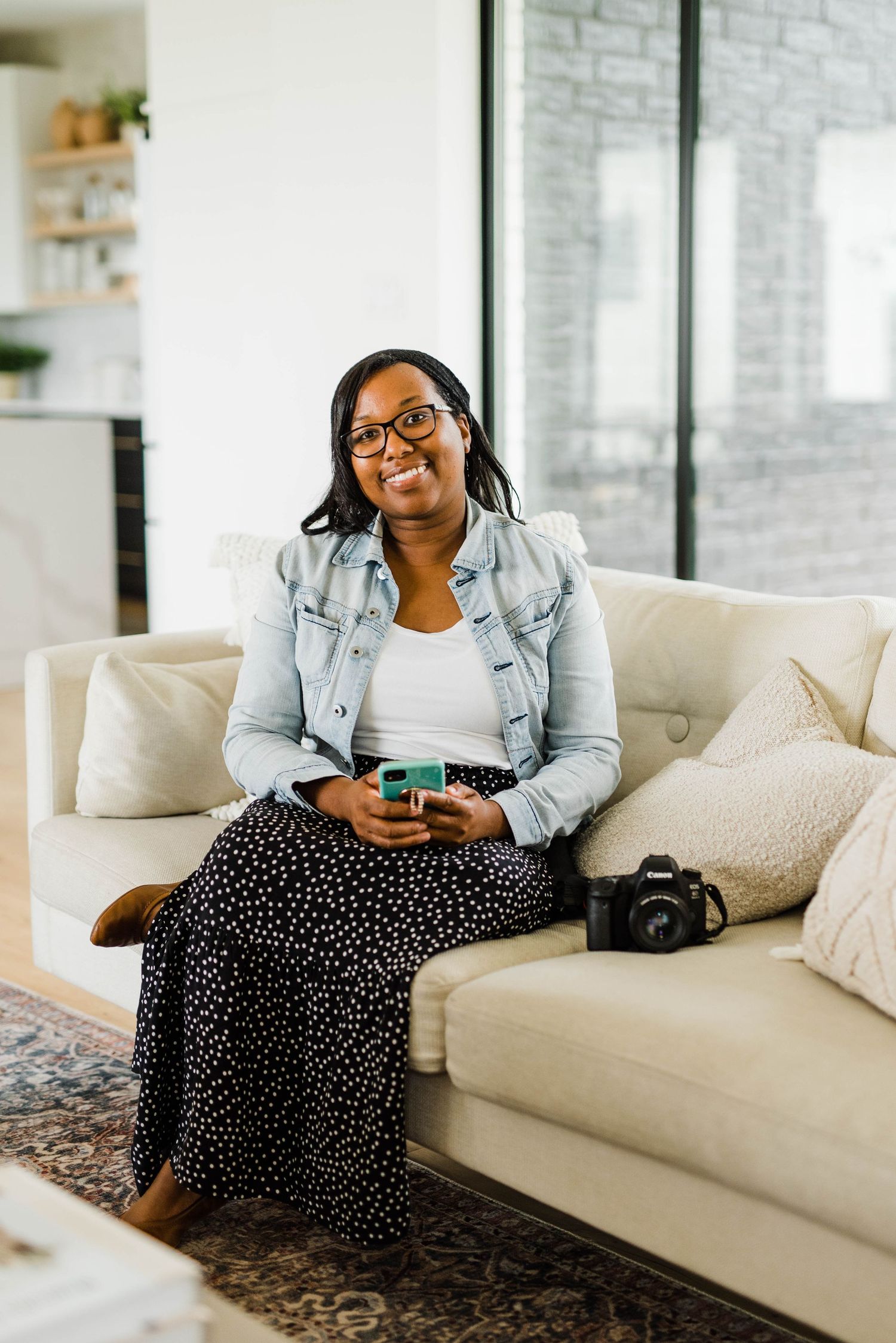 Woman relaxes on white couch wearing denim jacket and polka dot skirt while using phone.