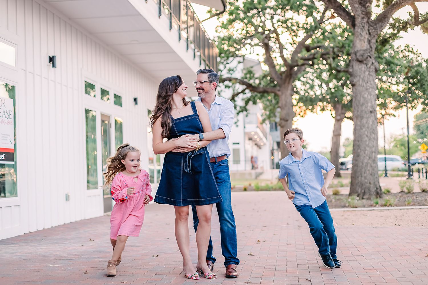 Family walking and playing along white building with brick pathway.