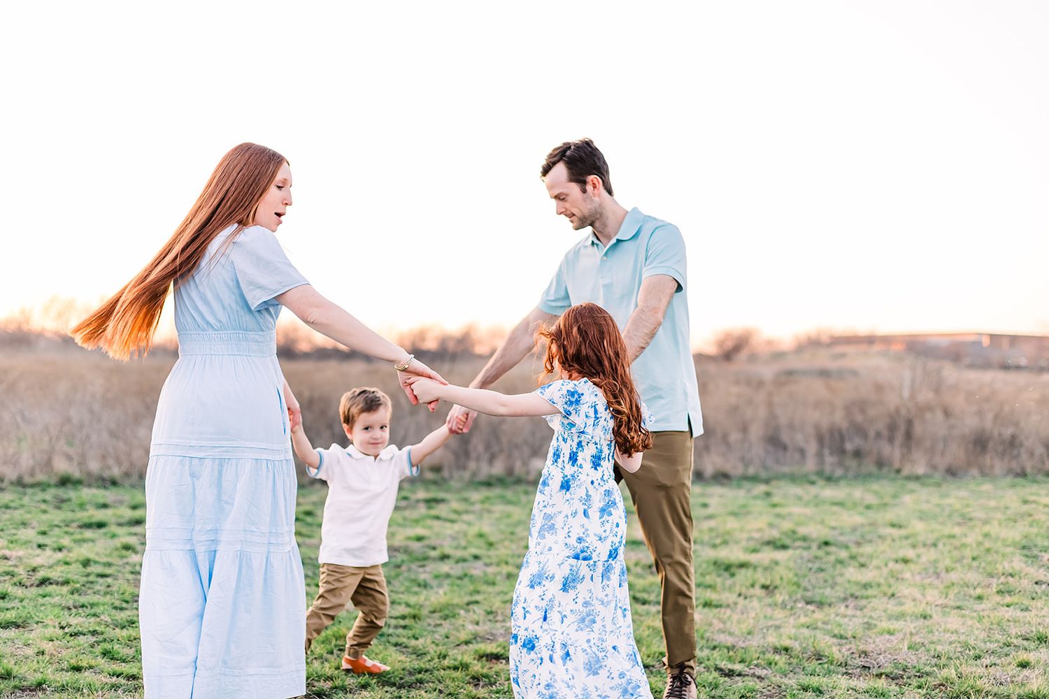 A family holds hands in a circle while playing together in a grassy field at sunset.